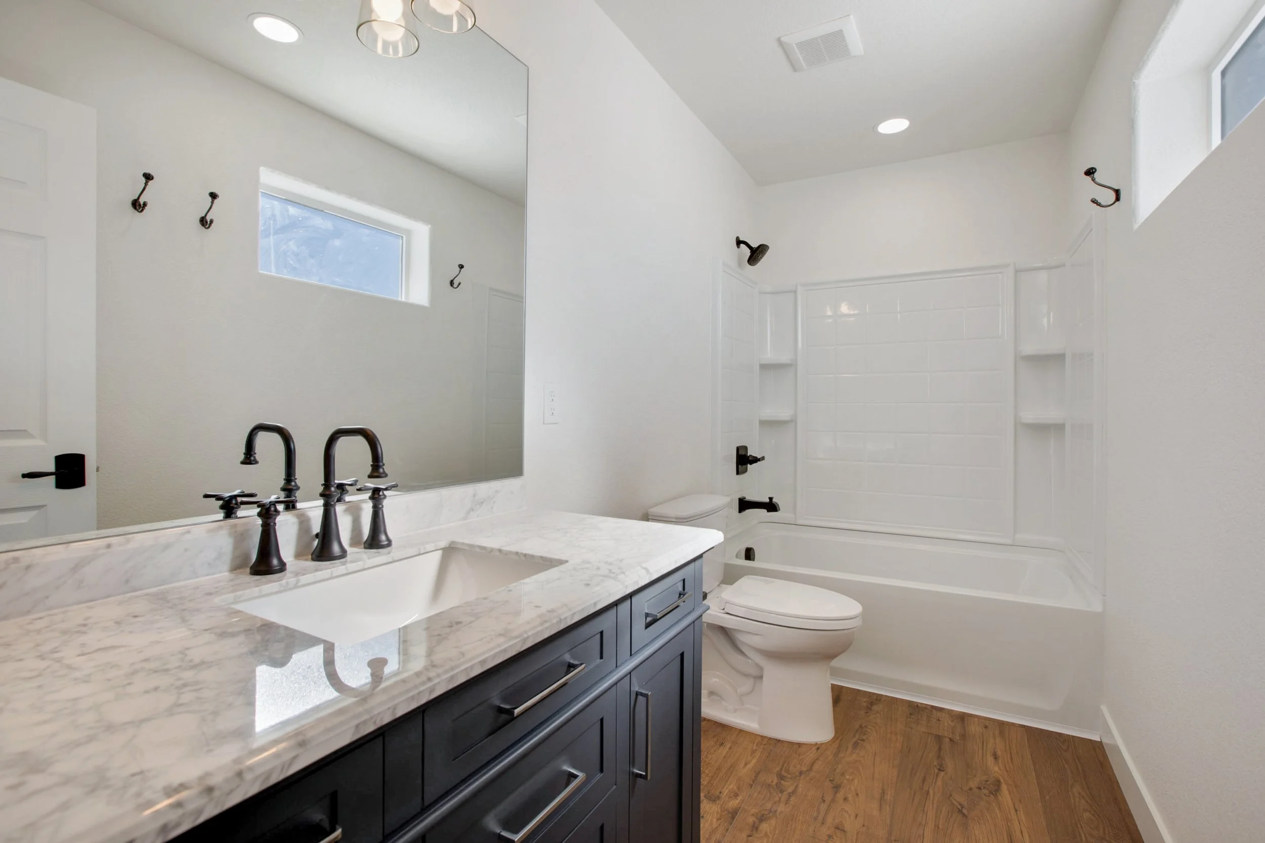 Bathroom with white walls, a large mirror above a marble countertop with a dark blue vanity, black fixtures, and a shower area with built-in shelves, a small window, and a toilet.