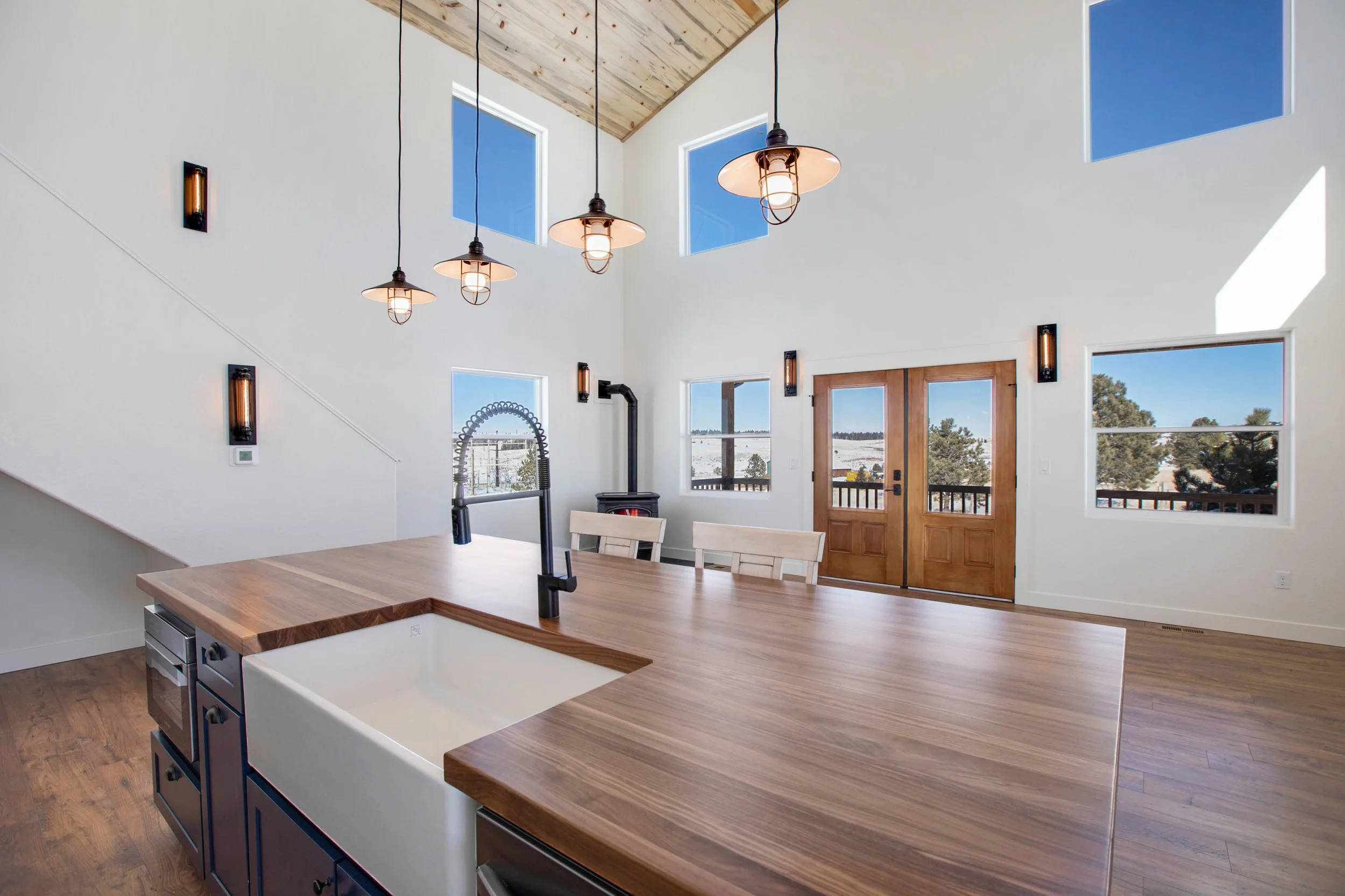 Modern kitchen interior with a large wooden island, black cabinets, white walls, multiple windows, and a ceiling with hanging pendant lights.