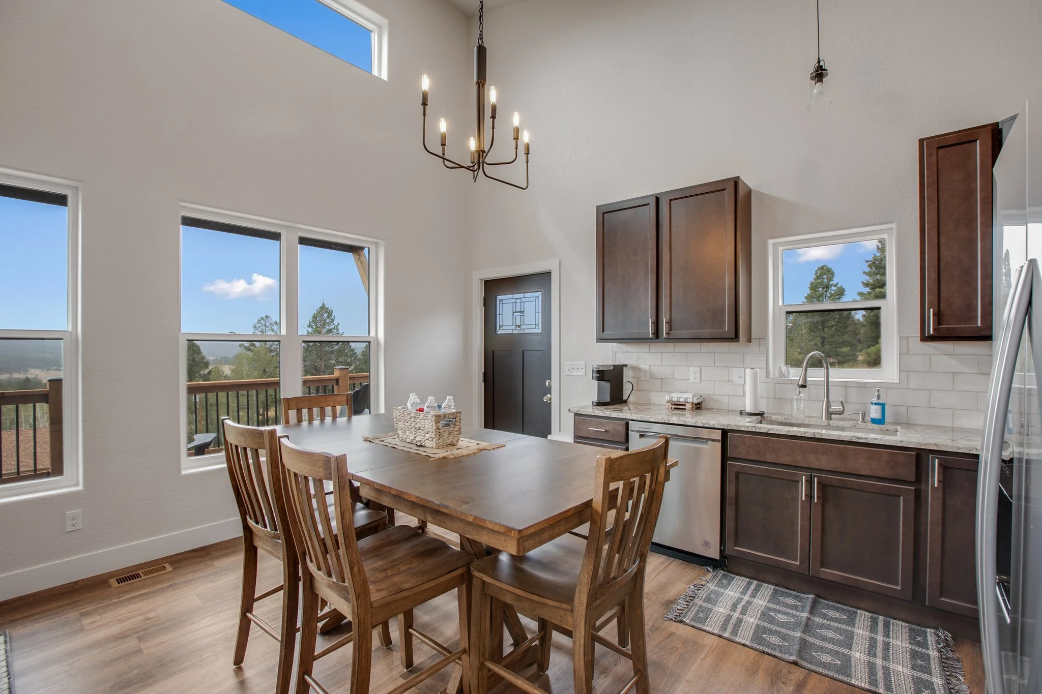A dining area with a wooden table, four chairs, and large windows showing a view of trees and a blue sky. The kitchen has dark wood cabinets, a gray and white marble countertop, and a stainless steel sink. A chandelier hangs from the ceiling, and a l