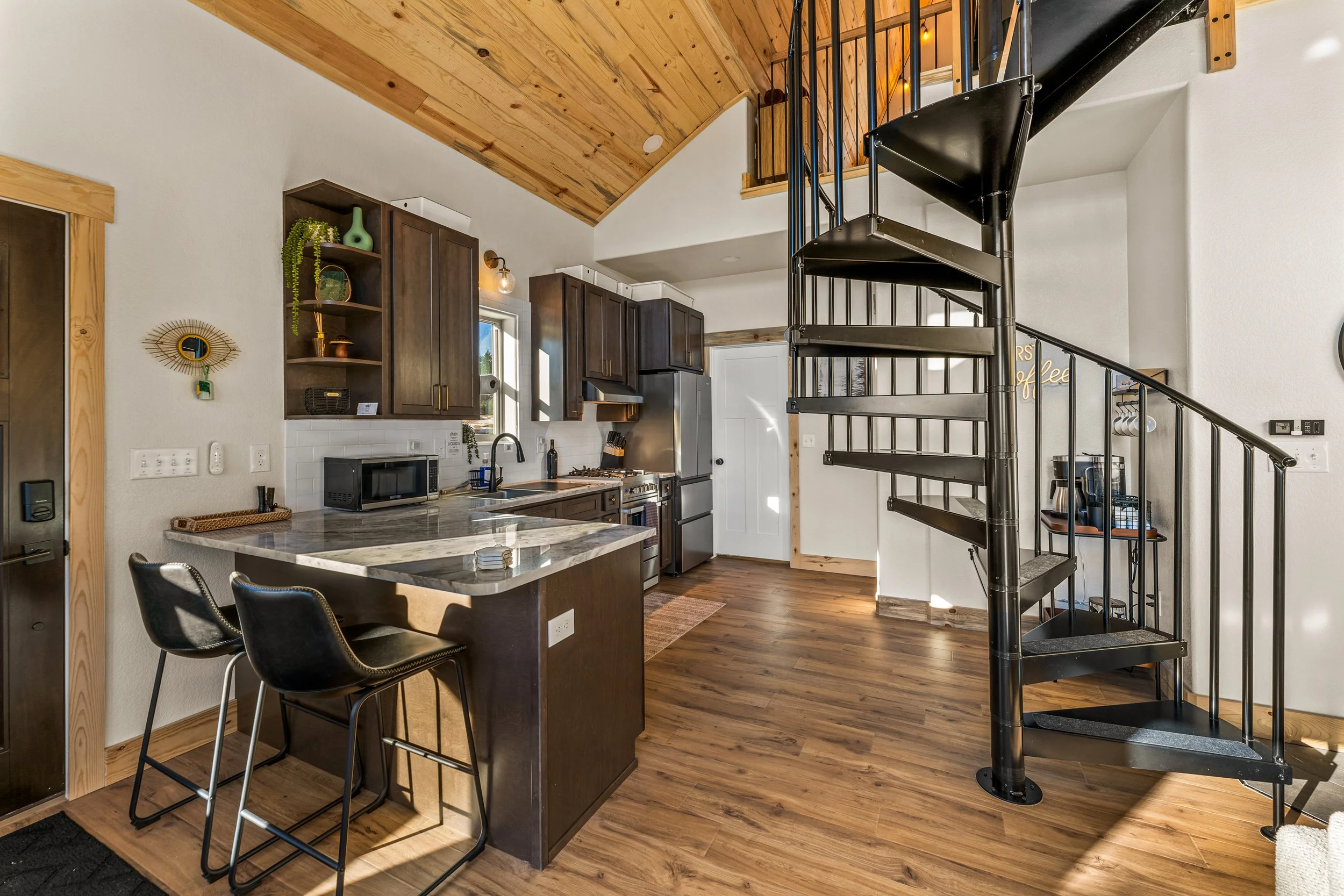 Interior view of a modern kitchen with dark wood cabinets, stainless steel appliances, a marble kitchen island, black chairs, wood flooring, a spiral staircase, and a wooden ceiling.