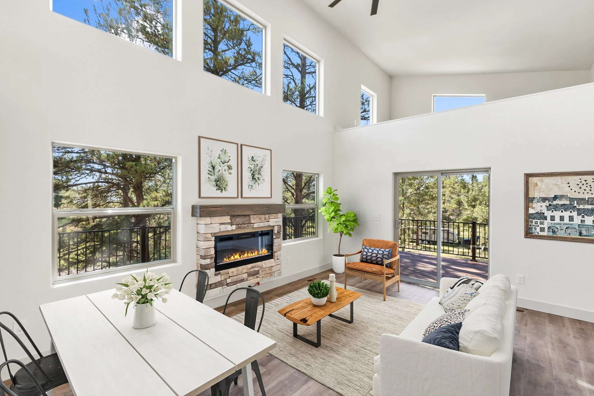 Interior of a modern living room with white walls, multiple windows showing trees outside, a stone fireplace with a fire burning, a white sofa, an orange armchair, a small wooden coffee table, framed botanical art, a potted plant, and sliding glass d