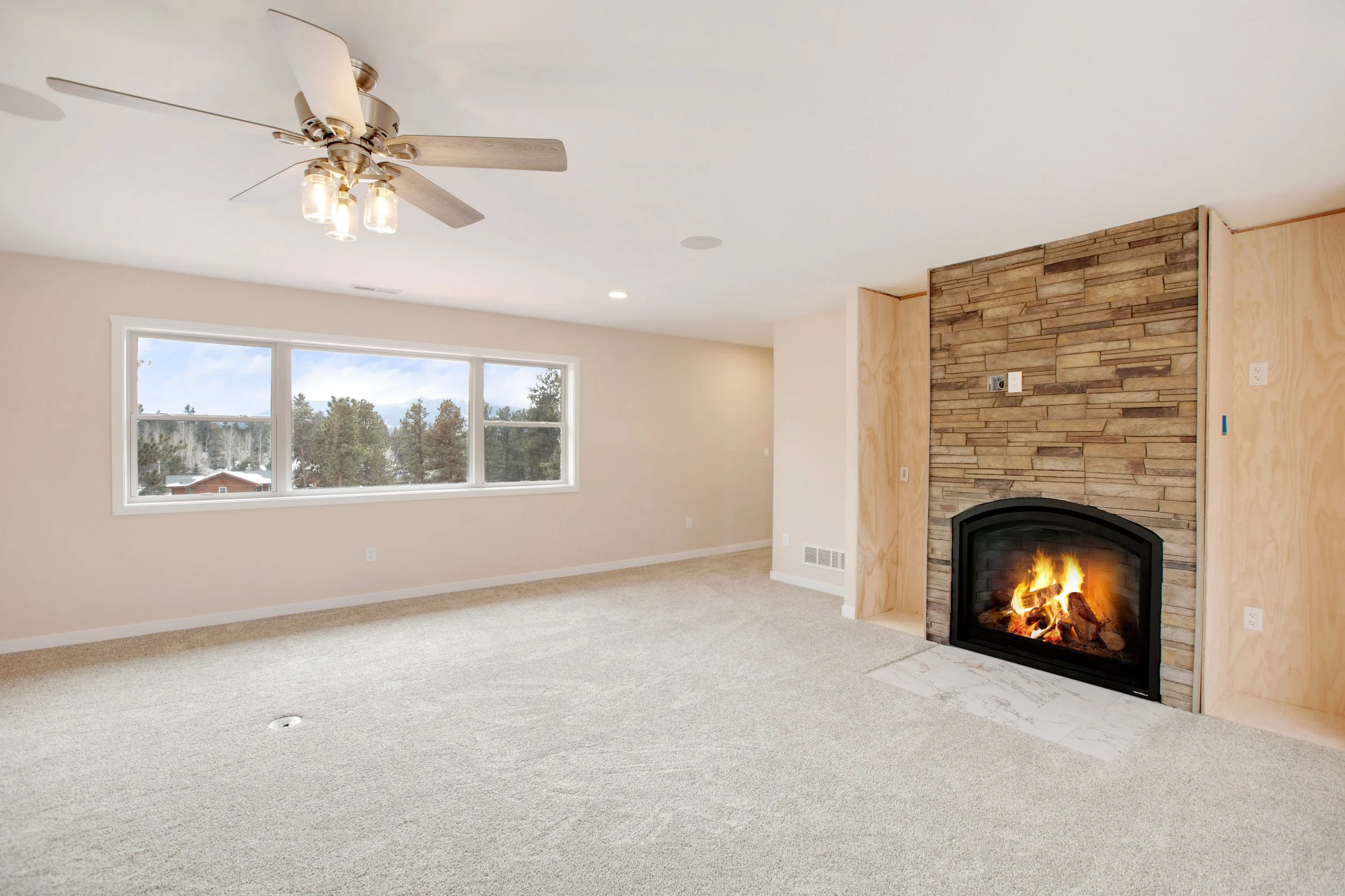A spacious living room with a large window showing trees and houses outside. The room features a stone fireplace with a fire burning, a ceiling fan with lights, and light-colored walls and carpet.