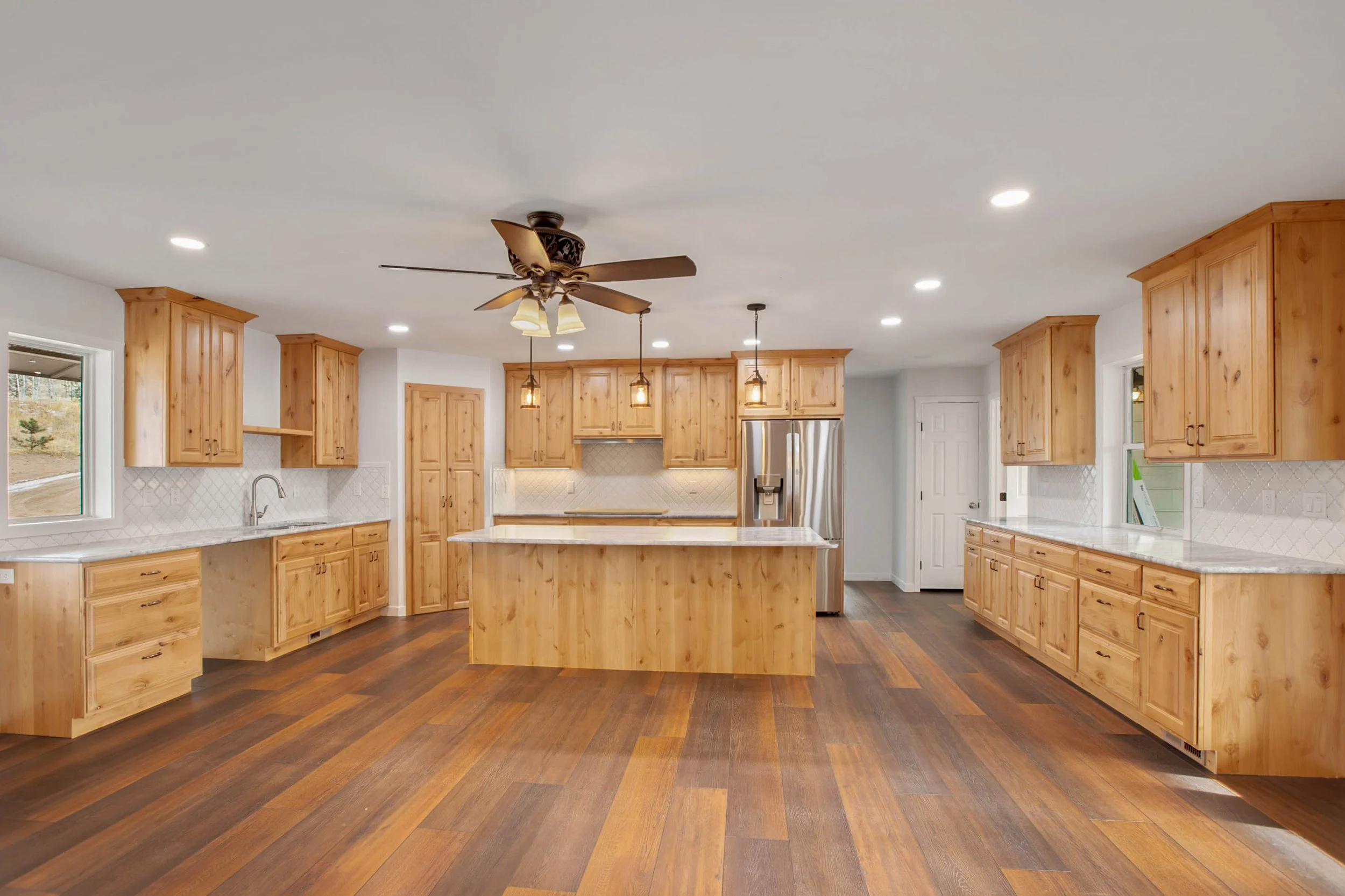 Bright kitchen with wooden cabinets, a white marble island, stainless steel refrigerator, and wood flooring, under a ceiling fan with lights.