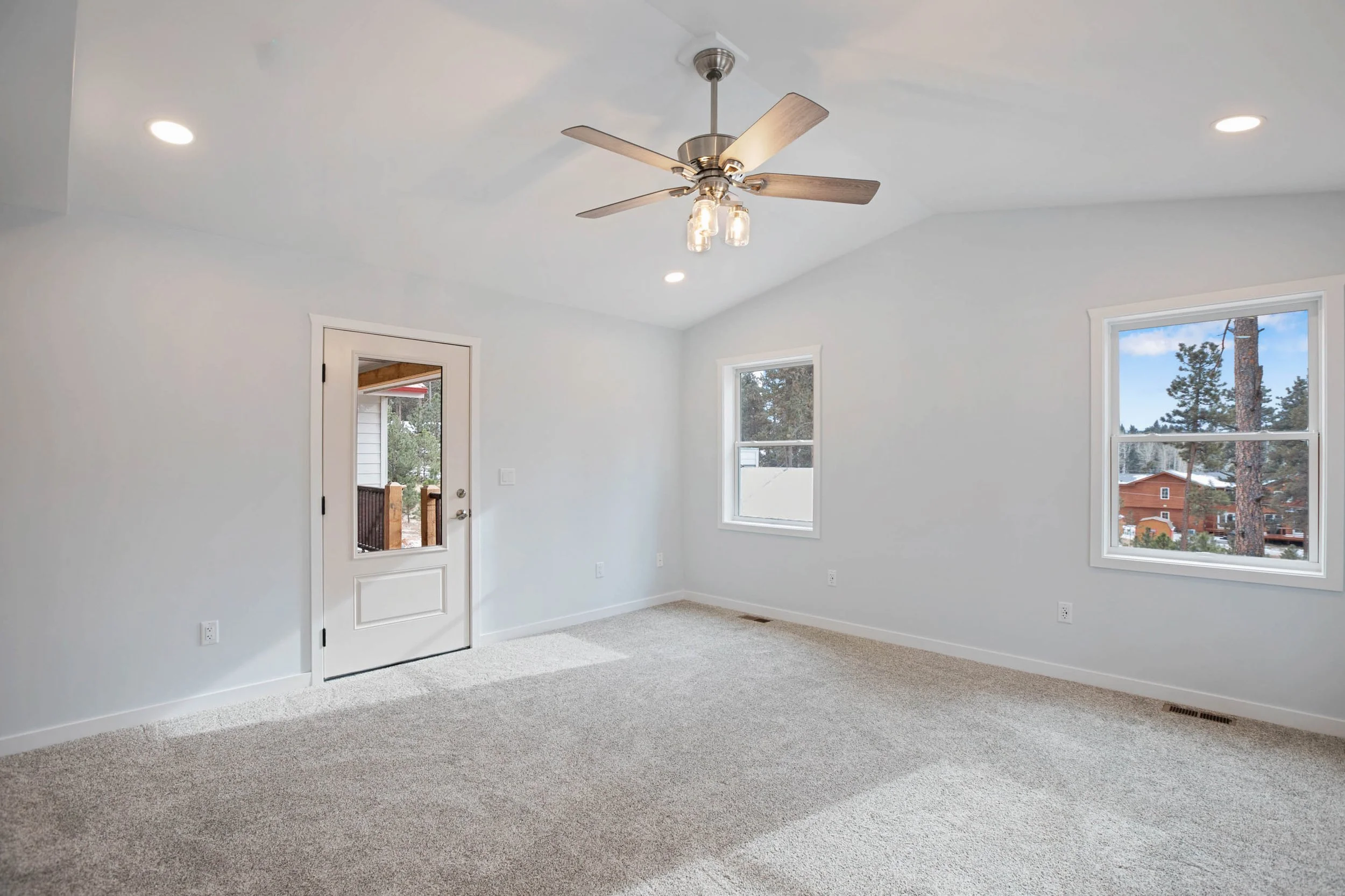 Empty room with white walls, beige carpet, windows showing a tree and a neighborhood, ceiling fan with lights, door leading outside, and recessed ceiling lights.