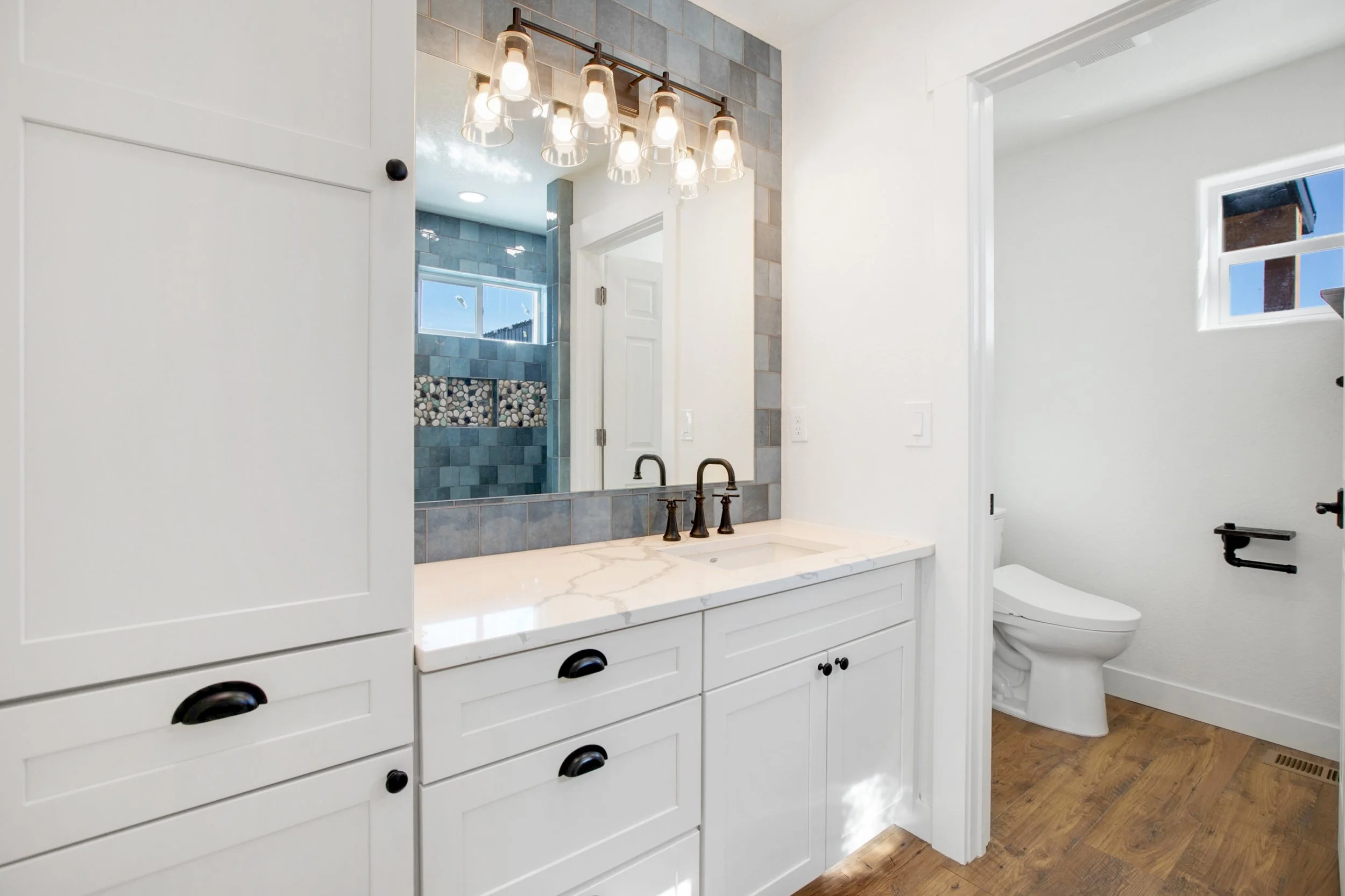 This image shows a modern bathroom with a white vanity, marble countertop, and black hardware. A large mirror above the sink is illuminated by a row of clear glass light fixtures. In the background, there is a toilet and a walk-in shower with blue an
