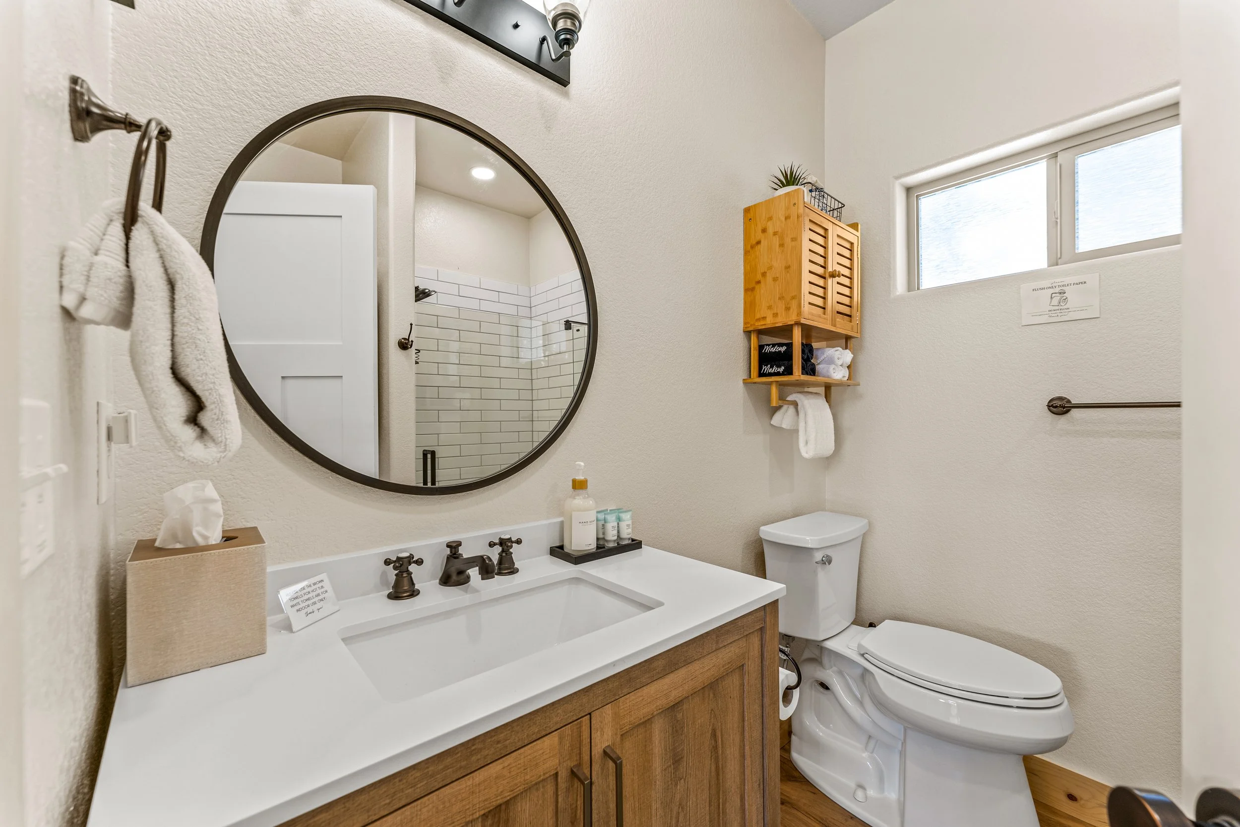 A bathroom with a white vanity and sink, black hardware, a large round mirror, a wooden cabinet on the wall, a window, and a toilet. There are towels, toiletries, and a tissue box visible.