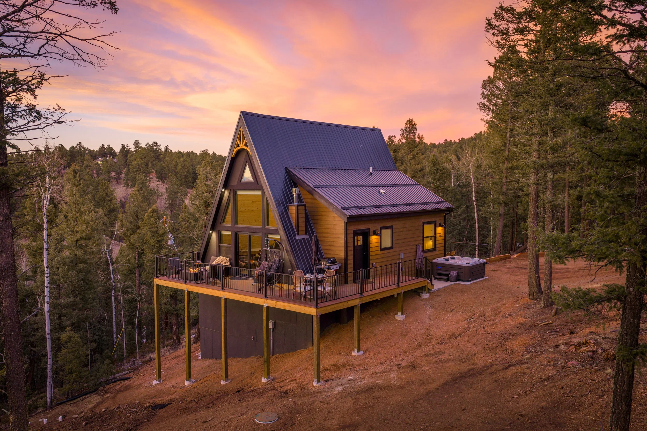 A-modern A-frame house with large windows on a hillside during sunset, surrounded by trees, with outdoor seating on a deck and a hot tub.