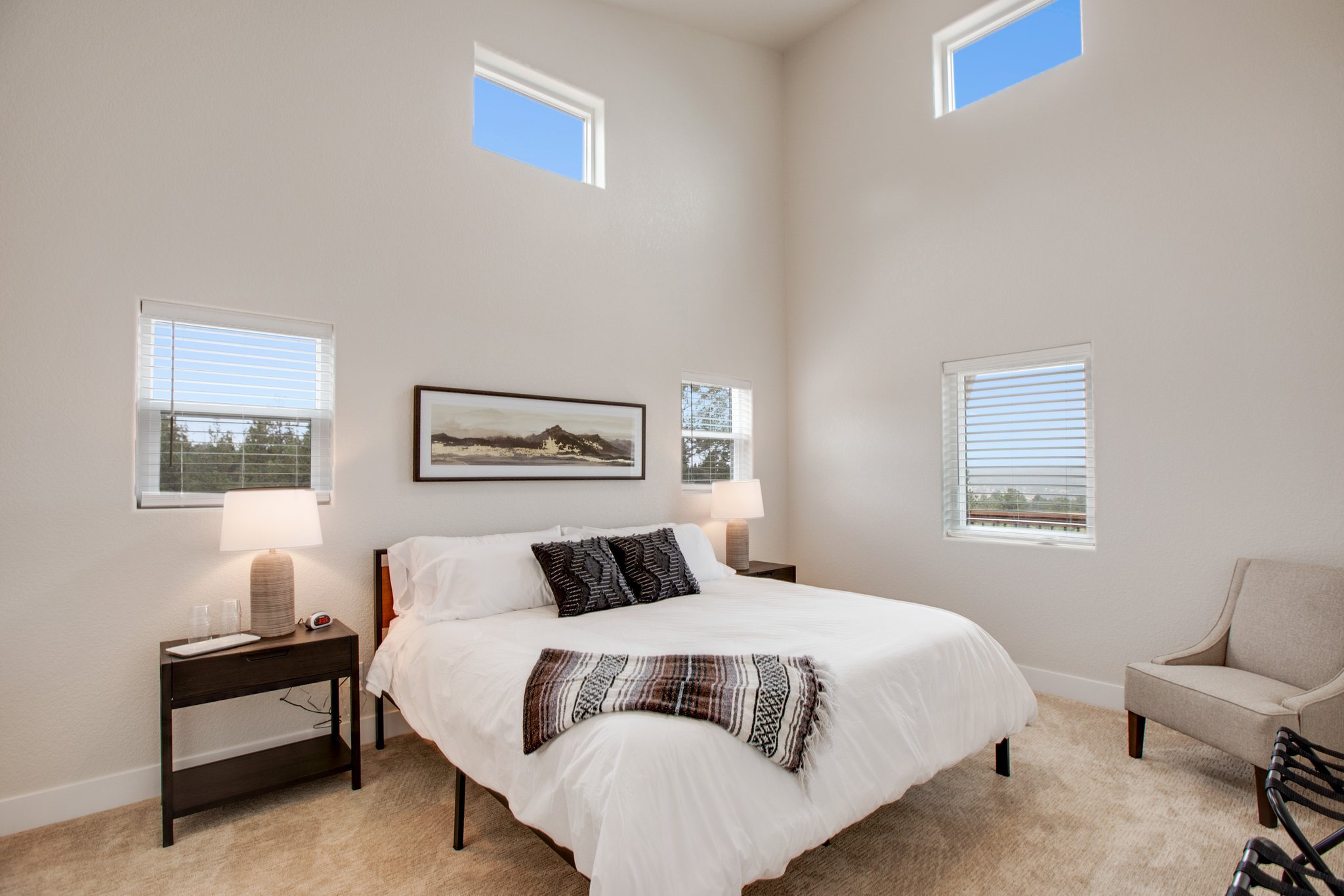 A bedroom with a bed, nightstands, lamps, artwork, and large windows letting in natural light.
