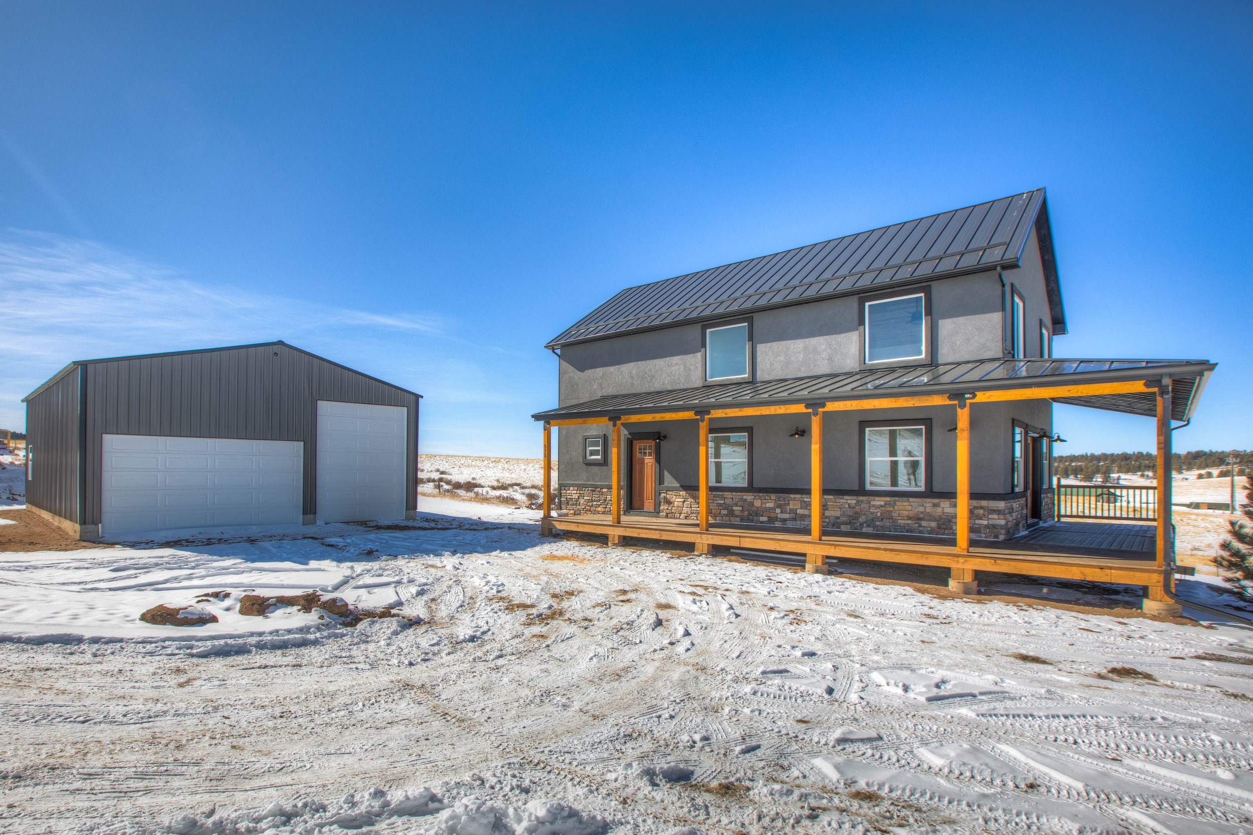 A house with a metal roof and a small porch, a detached garage, snow on the ground, and a bright blue sky.