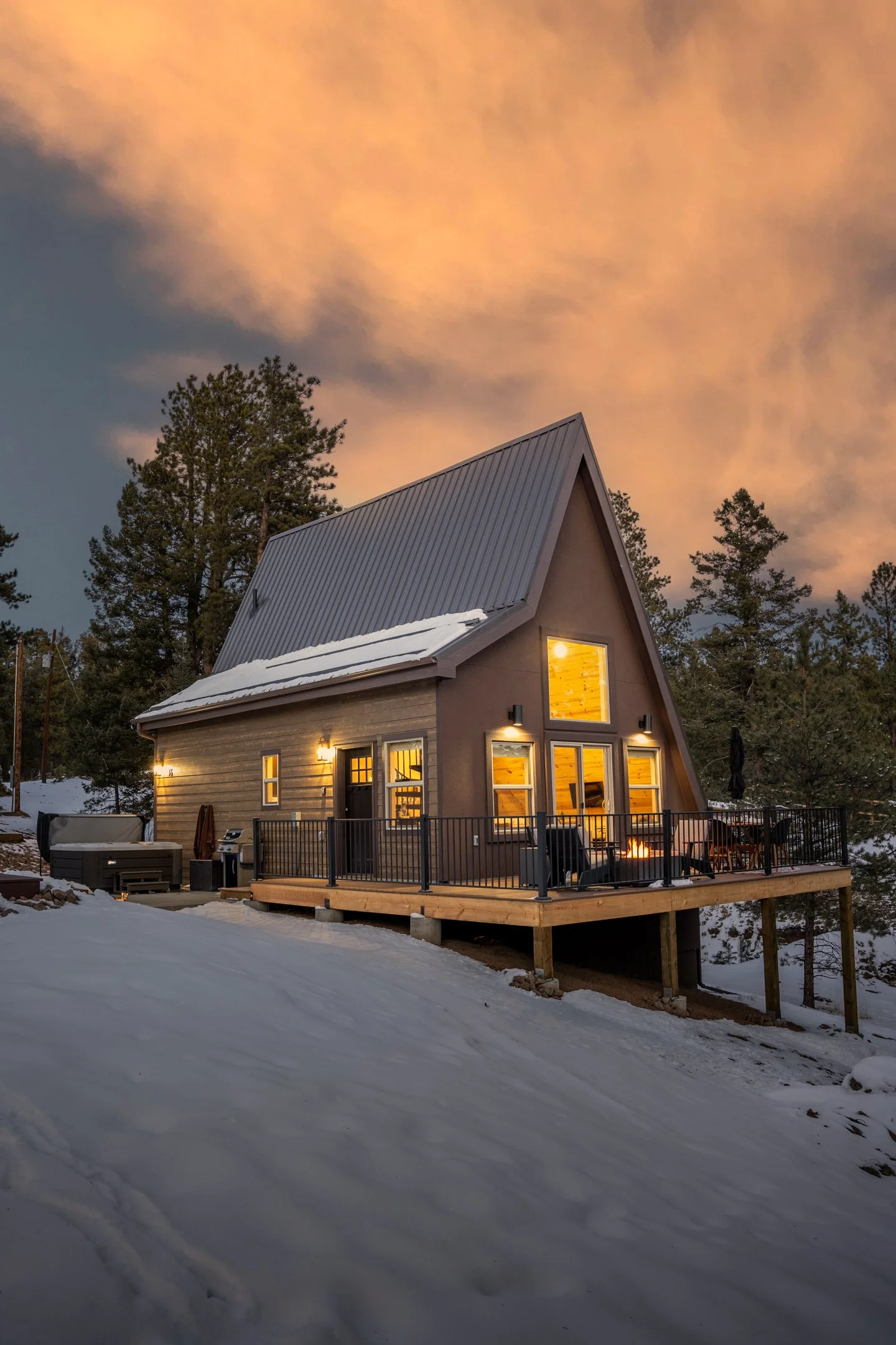 A modern house with a metal roof and large windows, illuminated from inside, situated in a snowy landscape surrounded by tall pine trees during dusk with a cloudy sky that has a pinkish hue.