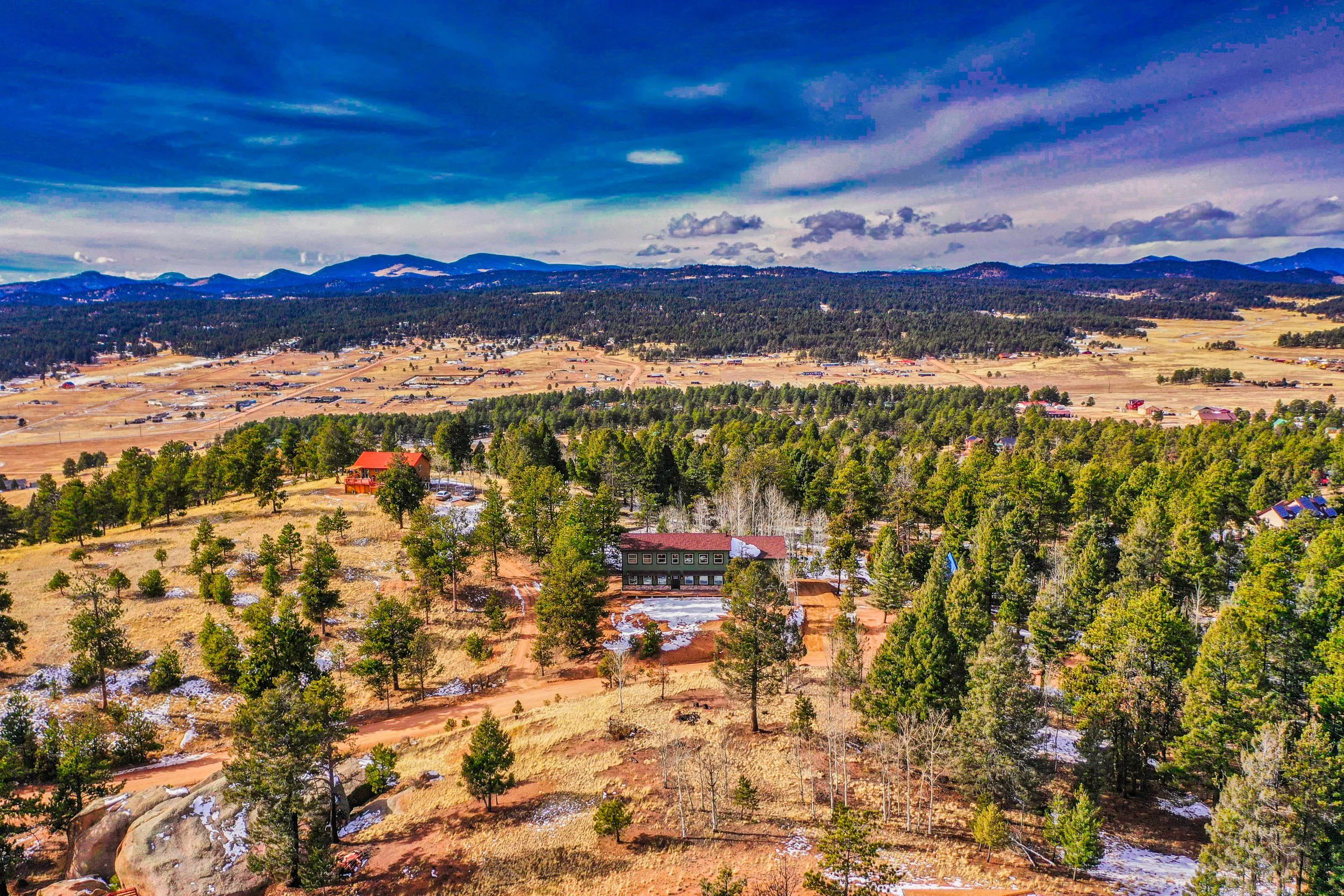 A scenic landscape of a mountain valley showing trees, grass fields, scattered houses, and a mountain range under a partly cloudy sky.