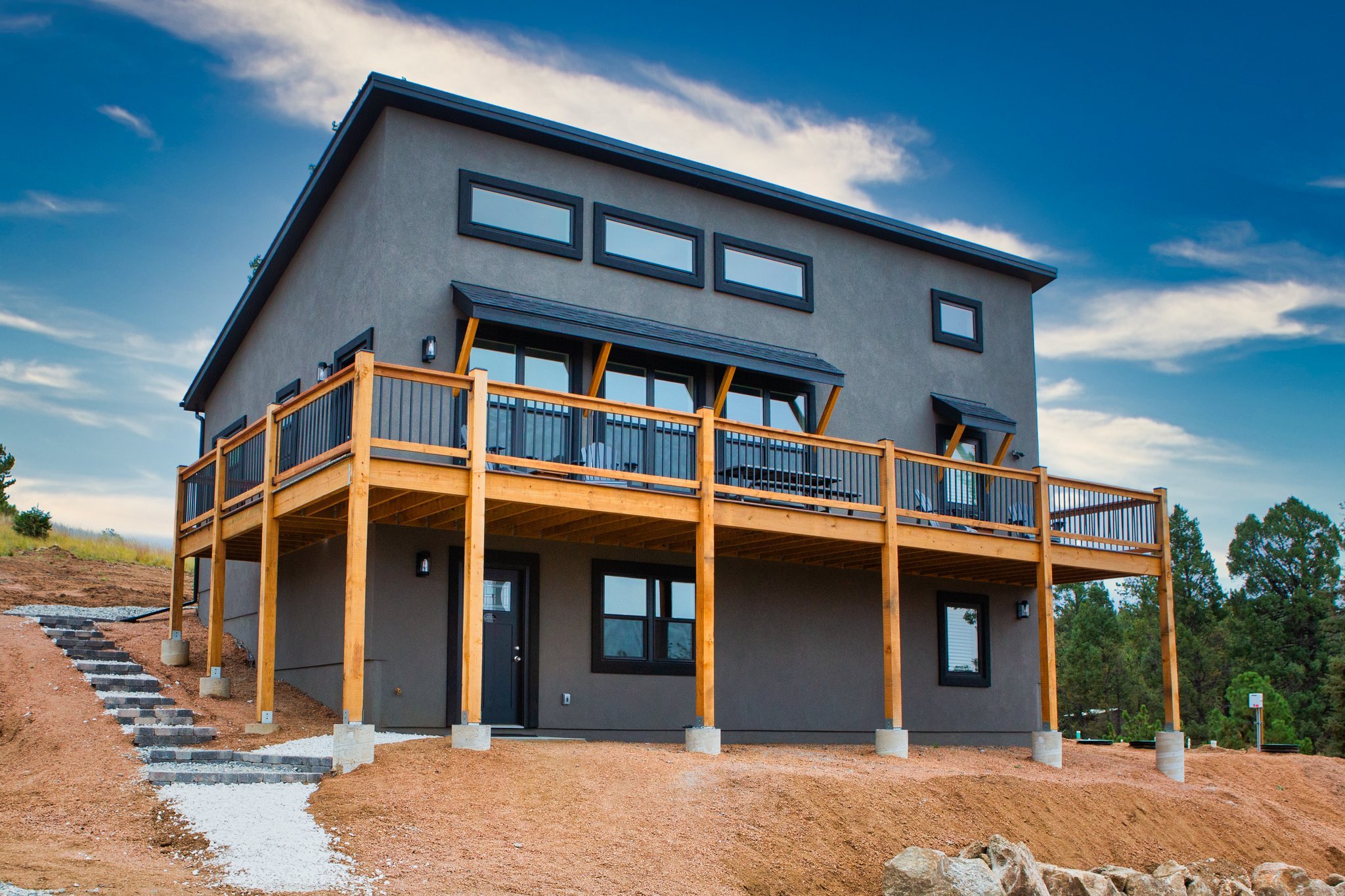 A modern, gray, three-story house with a balcony on the second floor, surrounded by dirt and construction materials, under a blue sky with clouds.