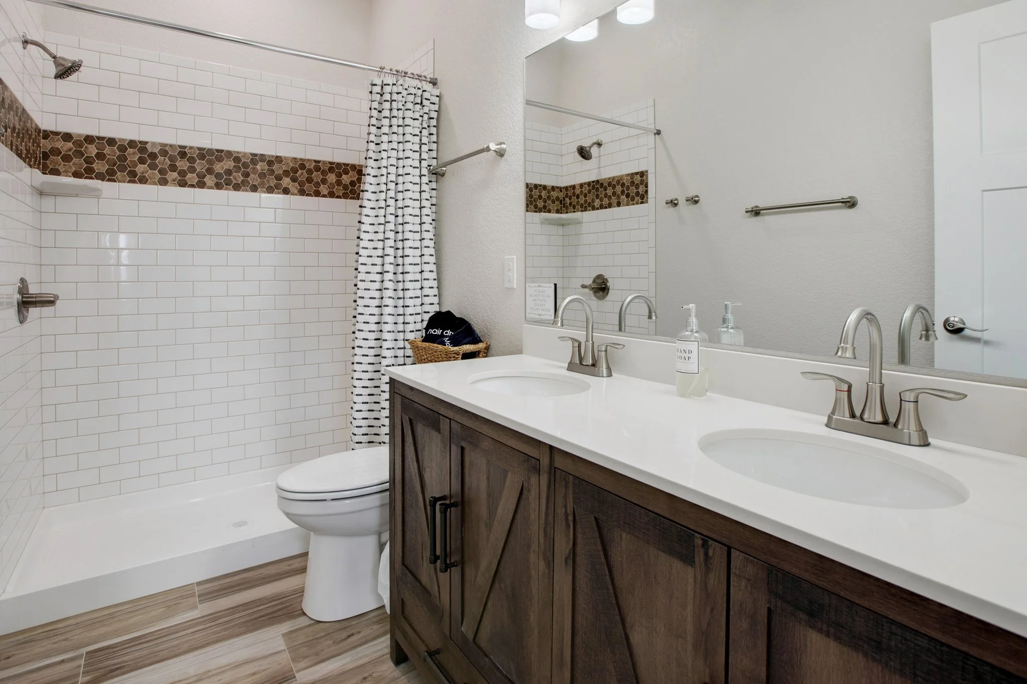 Modern bathroom with a walk-in shower, white tile walls with a decorative brown hexagon tile border, white vanity with double sinks, large mirror, and wood-look floor. Shower has a striped curtain, and bathroom accessories include hand soap on the co