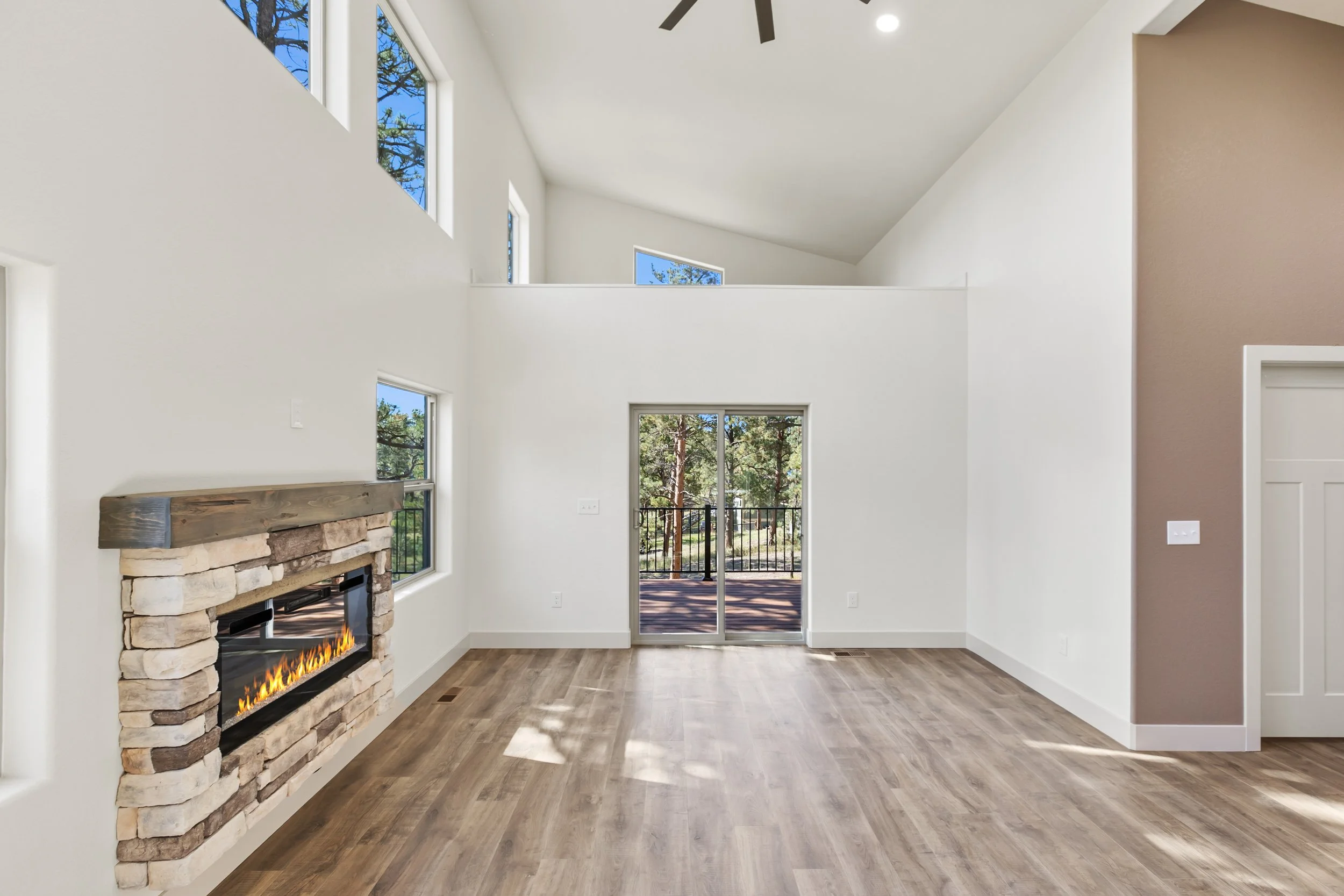 Interior of a modern living room with a stone fireplace, hardwood floors, large windows, and a view of trees outside, with a high ceiling and ceiling fan.
