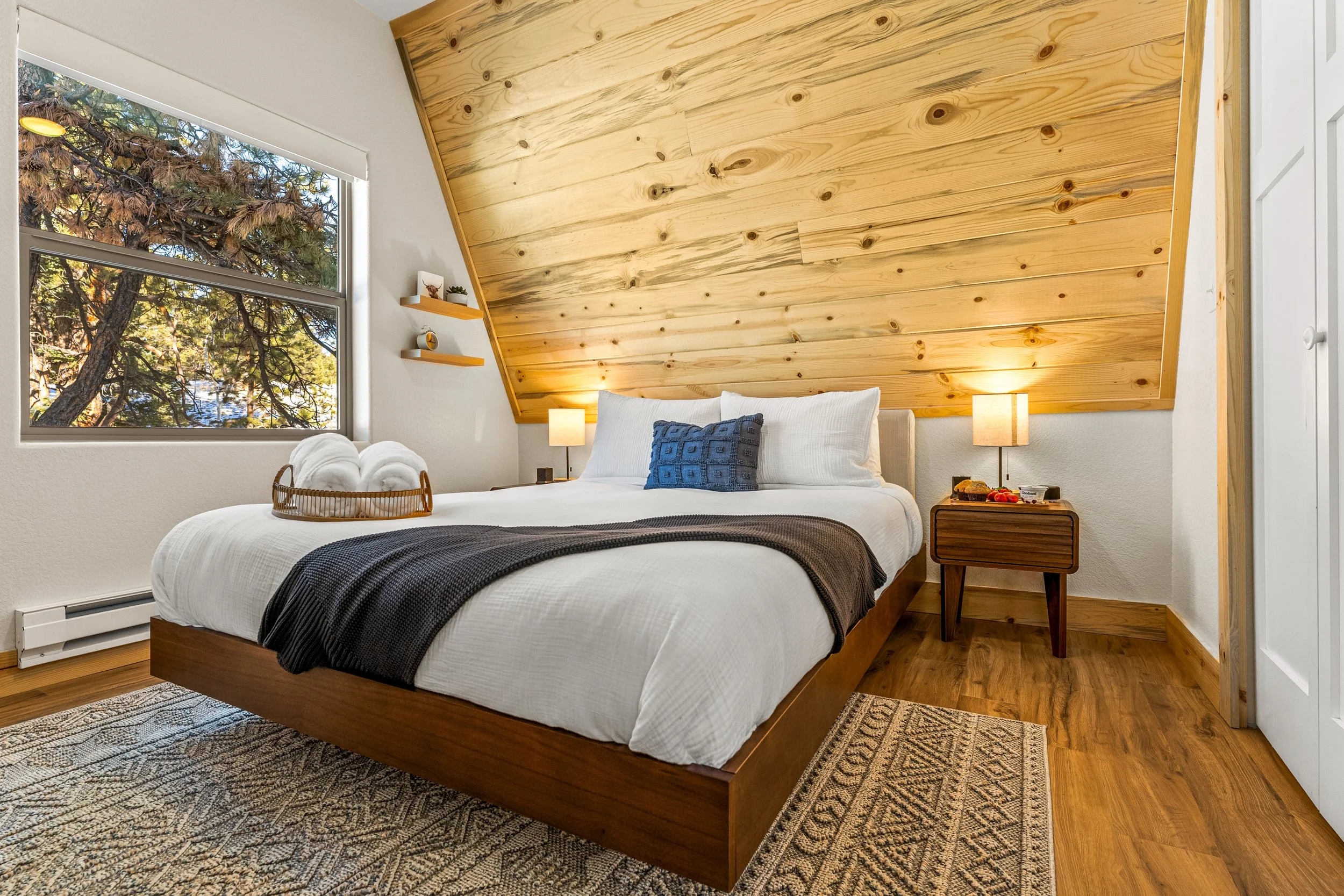 A bedroom with a wooden accent wall behind a bed, featuring white bedding and a decorative blue pillow. A window shows trees outside, and there are two small nightstands with lamps and decor items on either side of the bed. The room has a patterned r