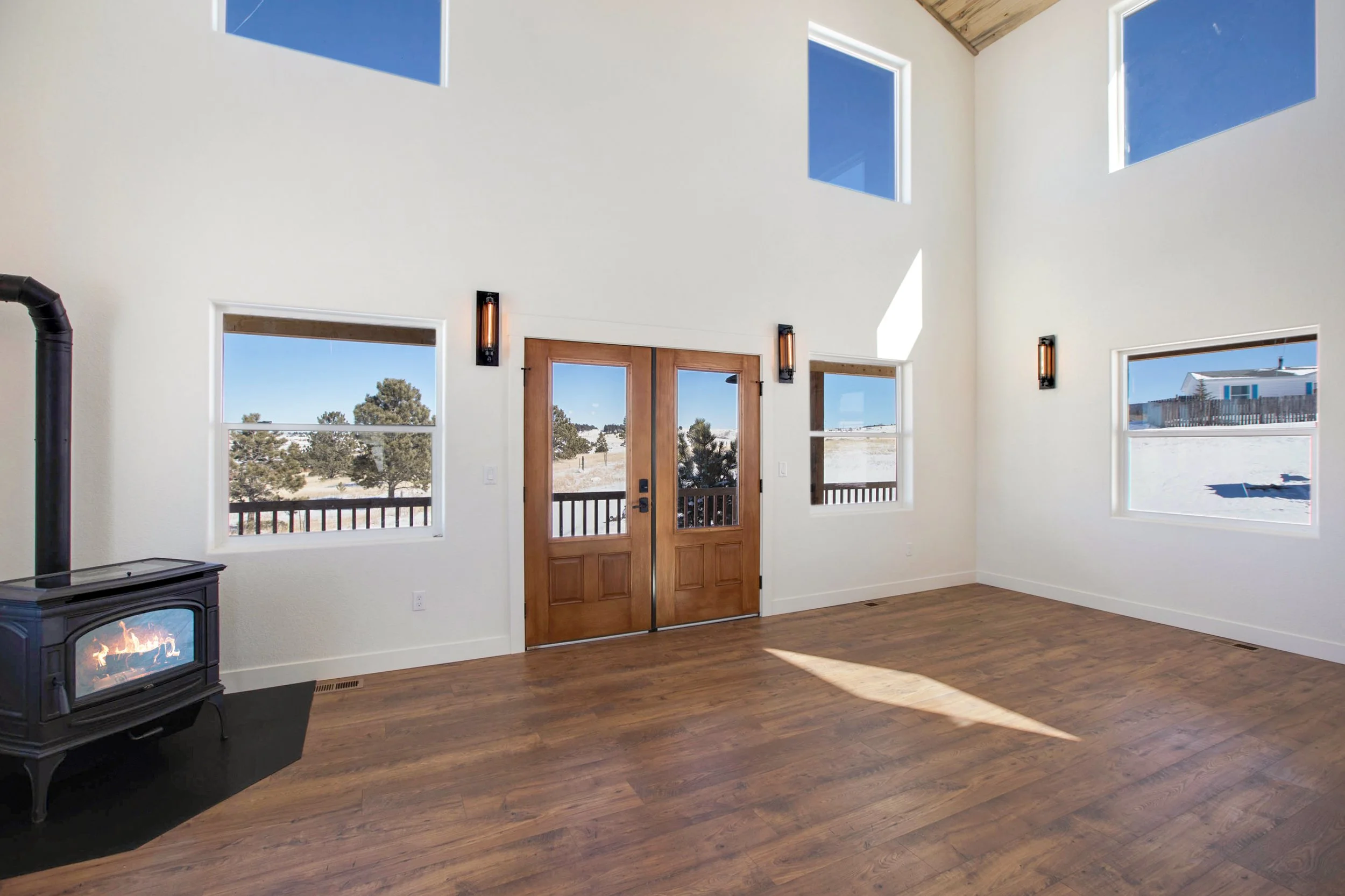 Interior of a room with white walls and large windows, featuring a wooden double door, hardwood flooring, a black stove with a fire inside, and wall-mounted light fixtures.