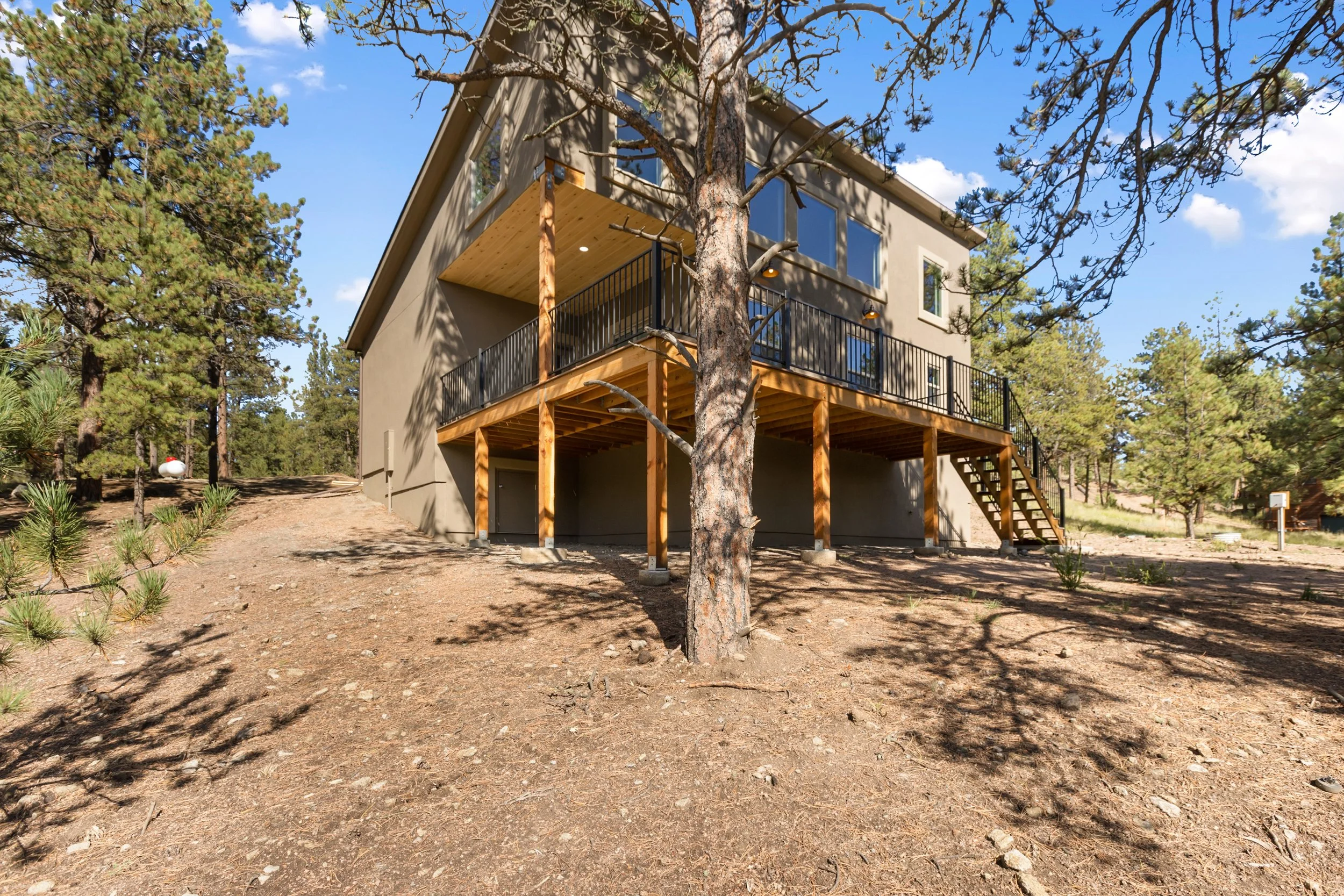 A two-story house under construction among trees, with a wooden deck and stairs leading to the ground, in a natural setting with dirt and pine trees.