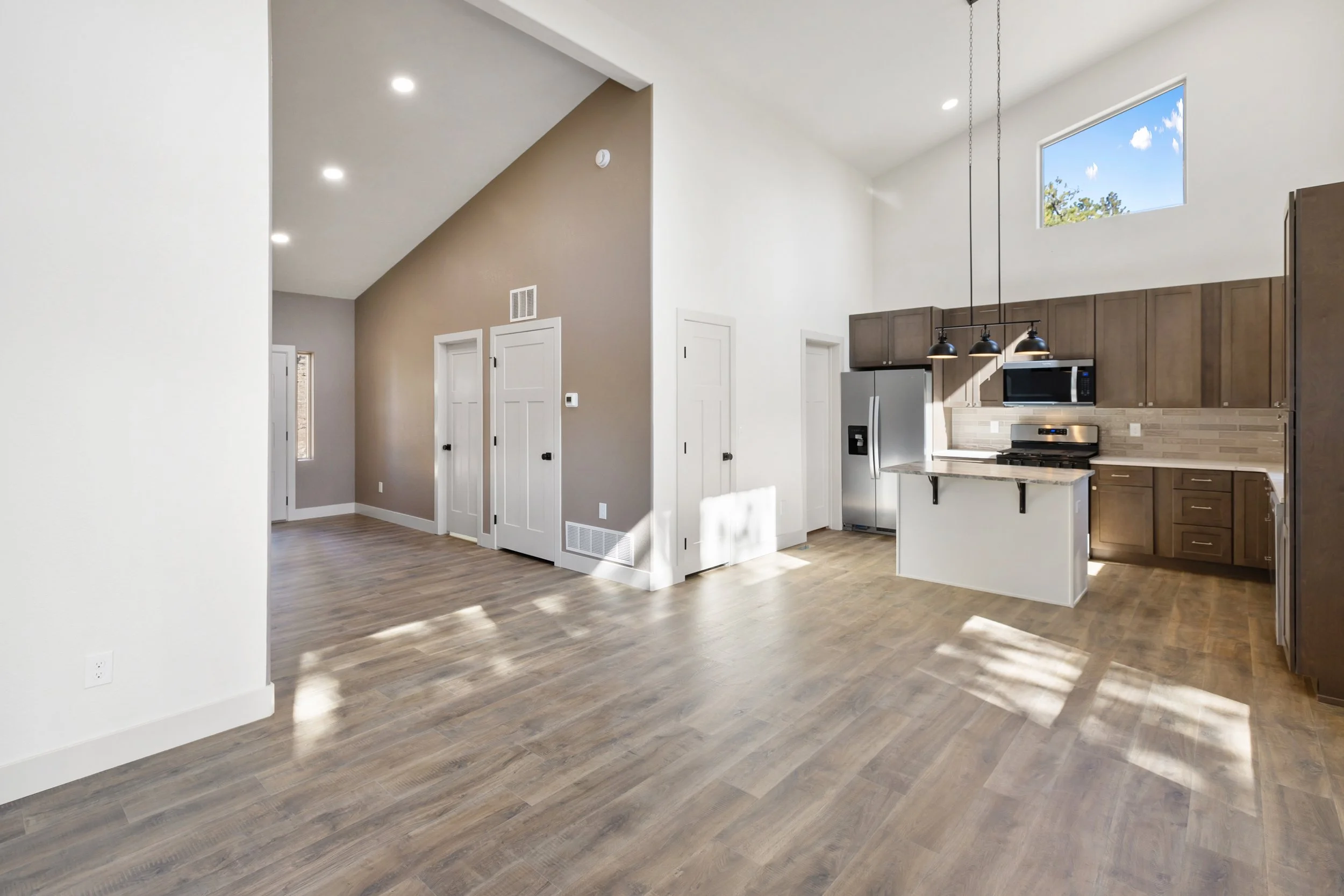 Empty living space with wood flooring, white and beige walls, kitchen area with stained wood cabinets, stainless steel refrigerator, microwave, oven, small island, and high ceilings with a large window.