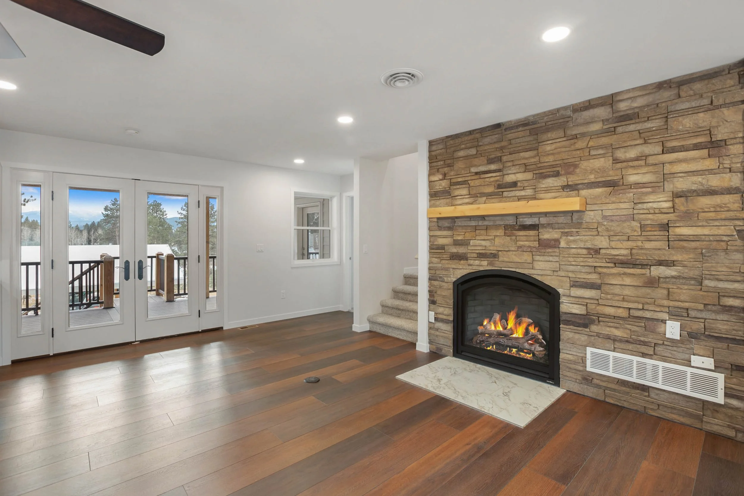 Living room with a fireplace, large windows, wooden floors, and a ceiling fan.