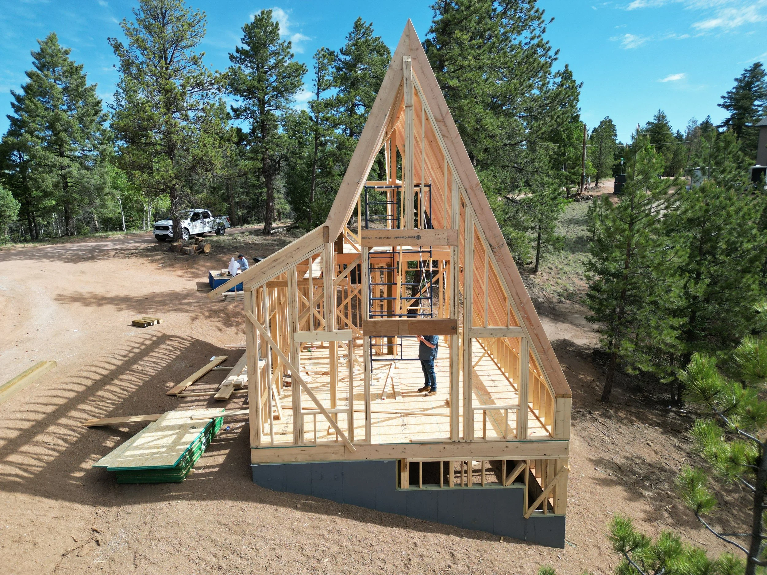 Wooden A-frame house under construction on a dirt lot surrounded by pine trees, with workers and vehicles nearby under a blue sky.