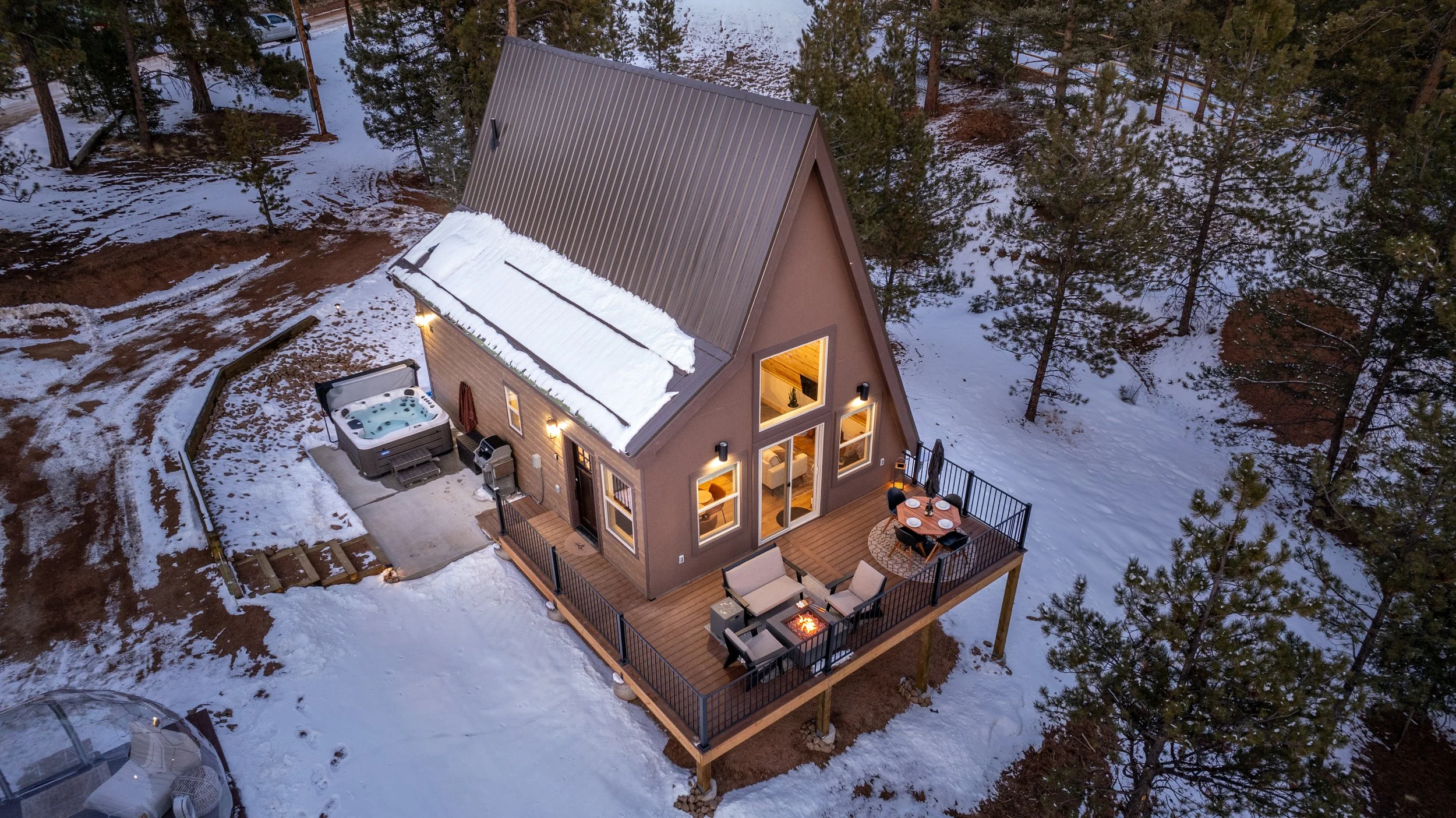 Aerial view of a small A-frame house with warm interior lights, surrounded by snow-covered ground and pine trees. The house has a wooden deck with outdoor furniture, a hot tub, and a fire pit.