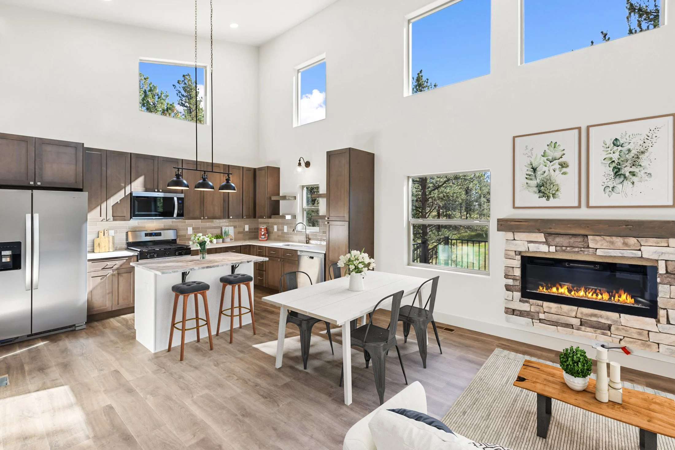 Bright living room with high ceilings, large windows showing trees and blue sky, a kitchen with dark wood cabinets, stainless steel appliances, a white island with bar stools, a white dining table with flowers, and a stone fireplace with artwork abov