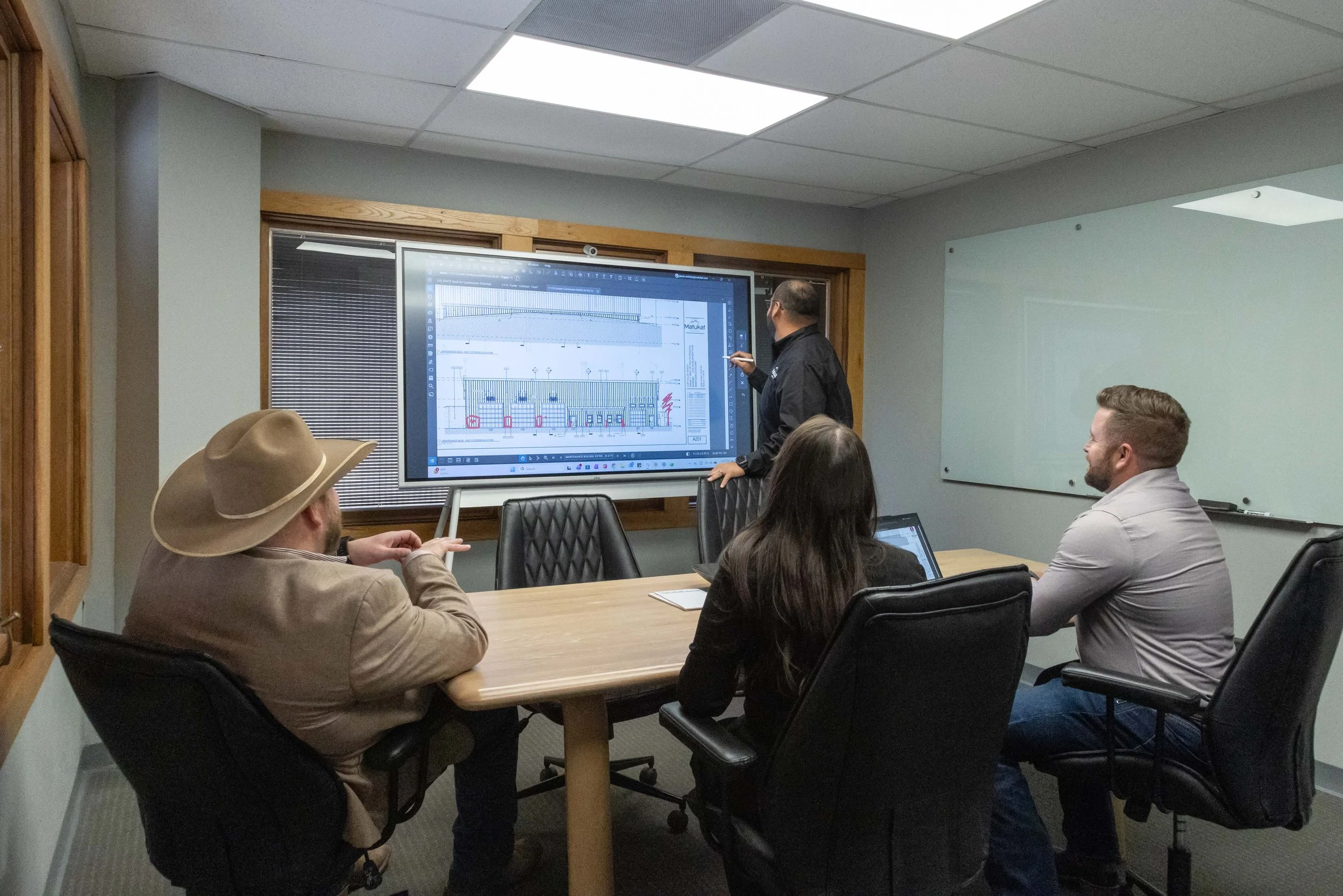 A group of people are sitting around a conference table in a meeting room. One person is standing and explaining a presentation on a large screen, which displays architectural or engineering drawings and graphs. The room has wood framing around the window and a whiteboard on the wall.