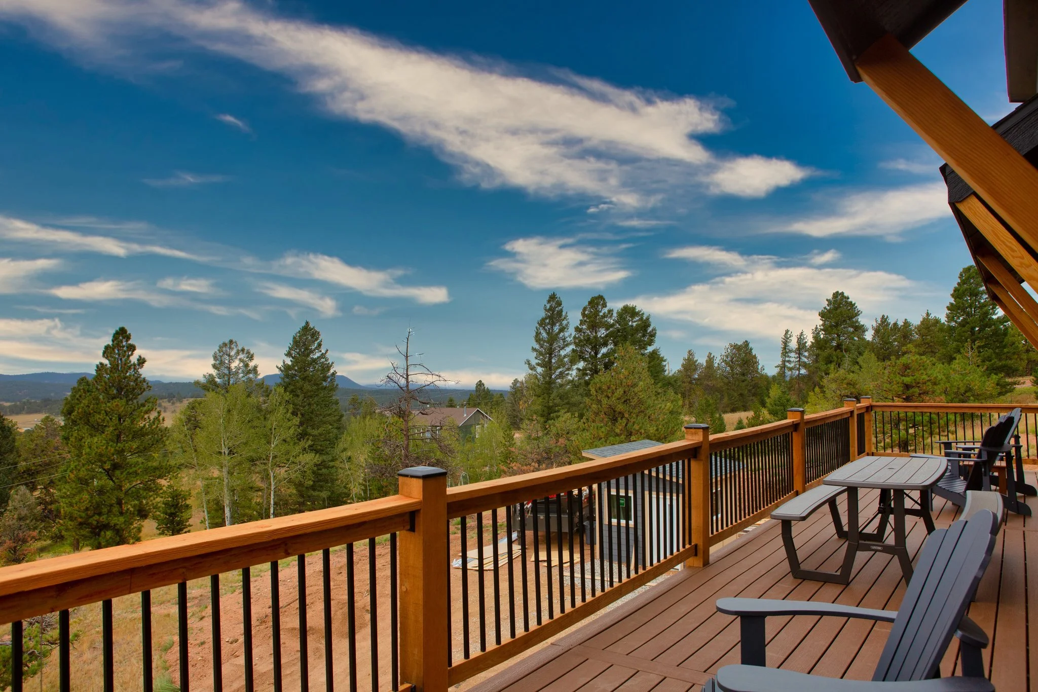 View from a wooden deck overlooking a forested landscape with tall pine trees and mountains in the distance under a partly cloudy blue sky.