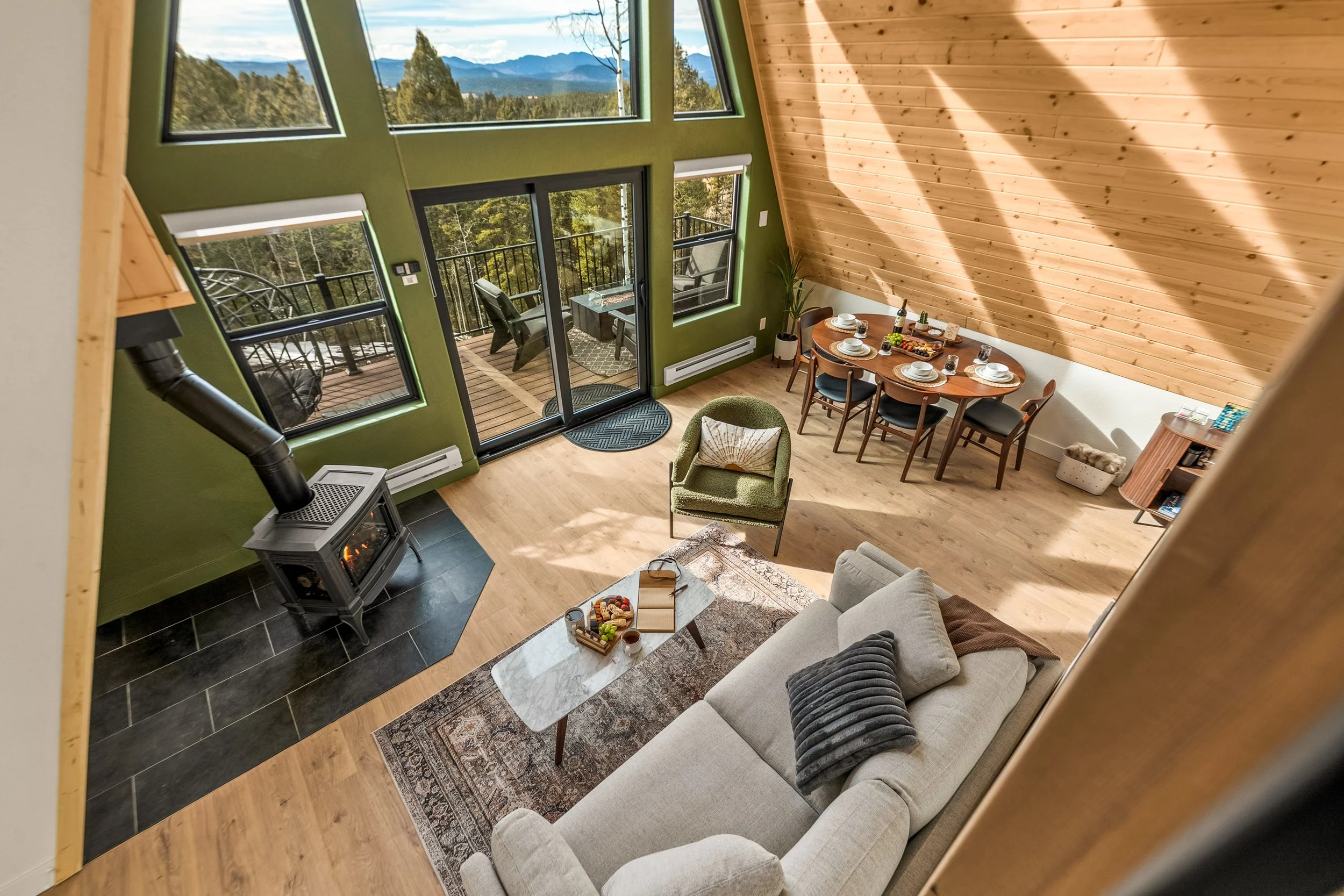 Living room with a beige sofa, a green armchair, and a marble-top coffee table with snacks, a tiled wood stove, a dining area with a wooden table and chairs set for a meal, and a sliding glass door leading to a balcony with outdoor furniture, overloo