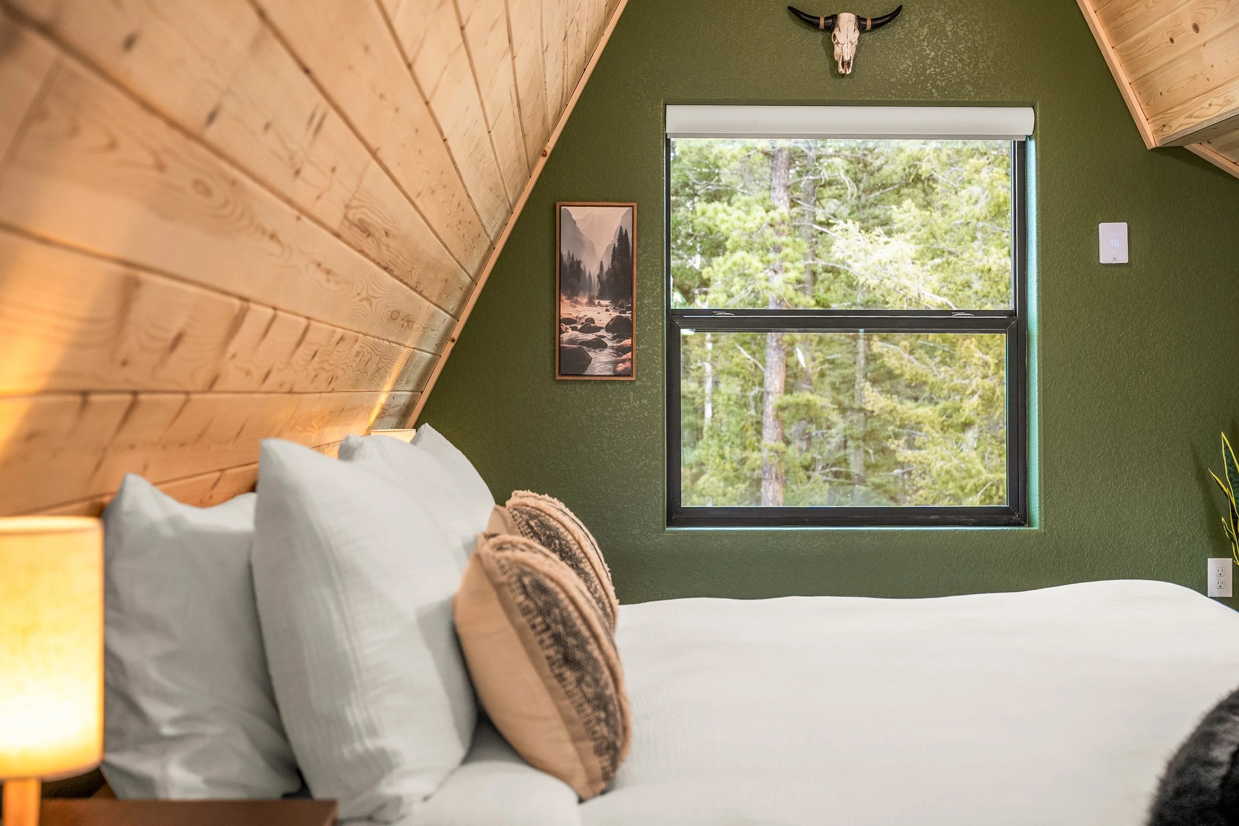 A bedroom with a green textured wall and a window showing trees outside. There is a framed mountain landscape picture on the wall, a bed with white pillows, a beige blanket, and a yellow bedside lamp. A decorative horned skull is mounted on the wall 