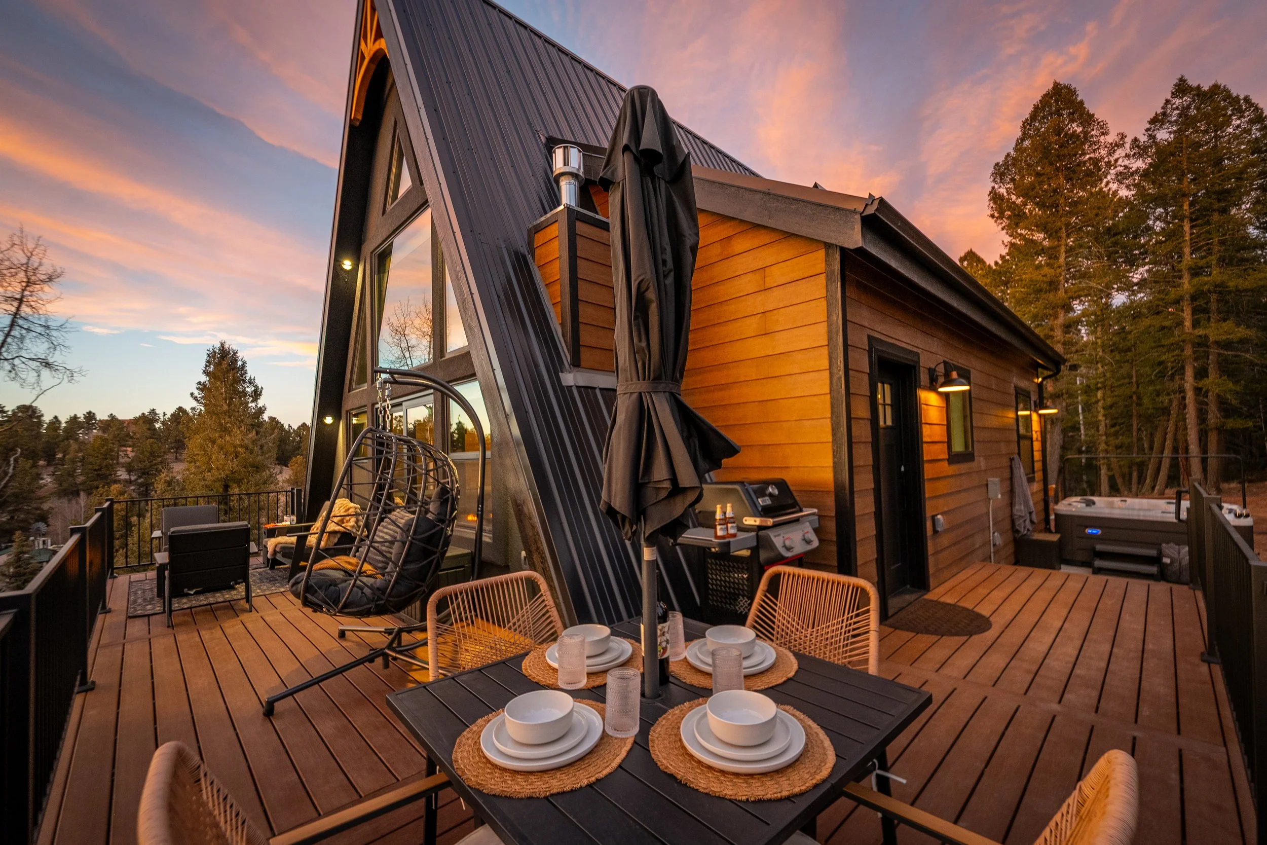 A wooden deck outdoor patio with a dining table set for six, featuring bowls, glasses, and placemats, surrounded by chairs, with a sitting area, barbecue grill, and tall trees in the background during sunset.
