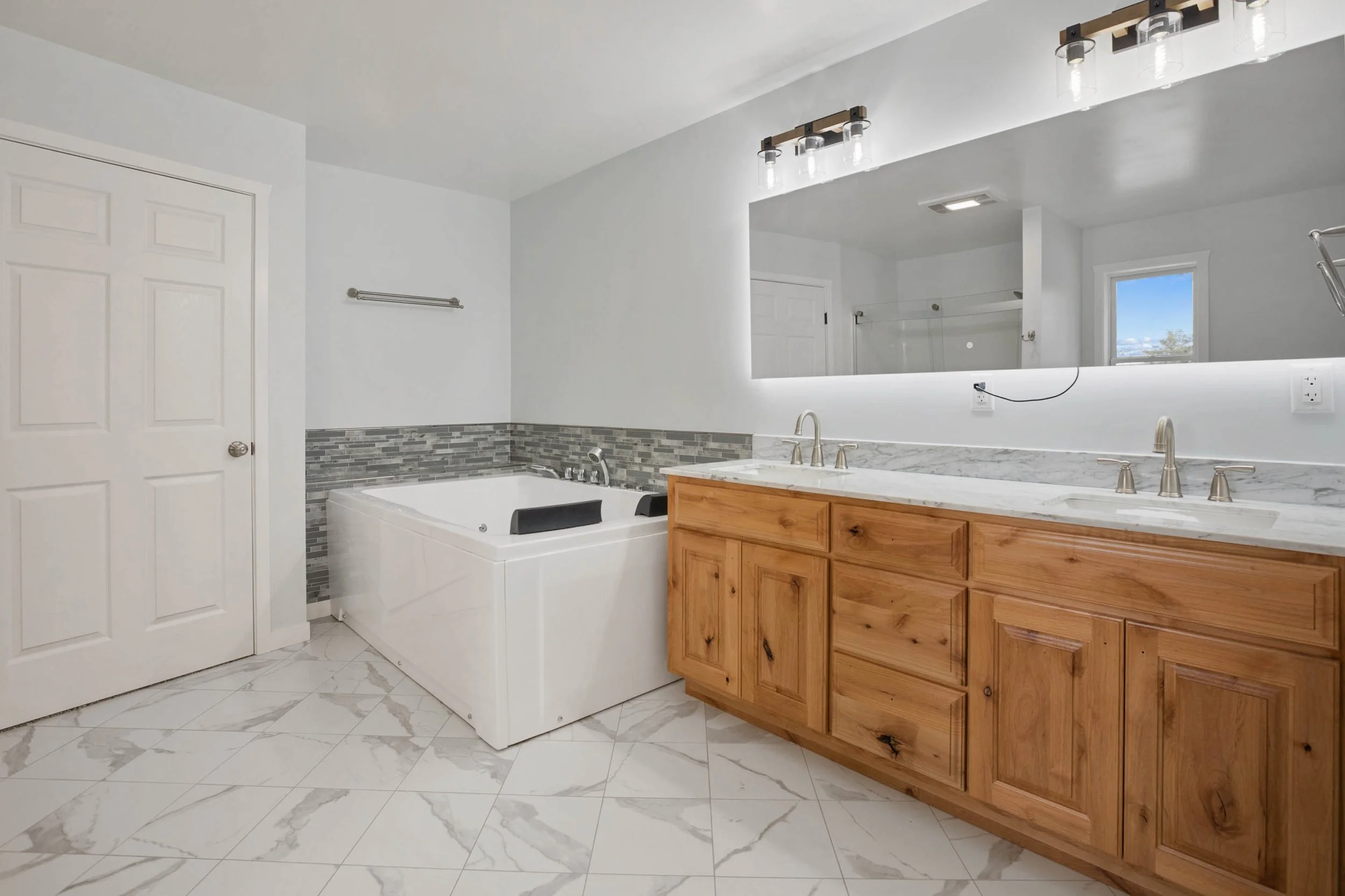 A modern bathroom with a large mirror, double sink vanity with wooden cabinets, a Jacuzzi bathtub, tiled walls, and a window with a view of the outdoors.