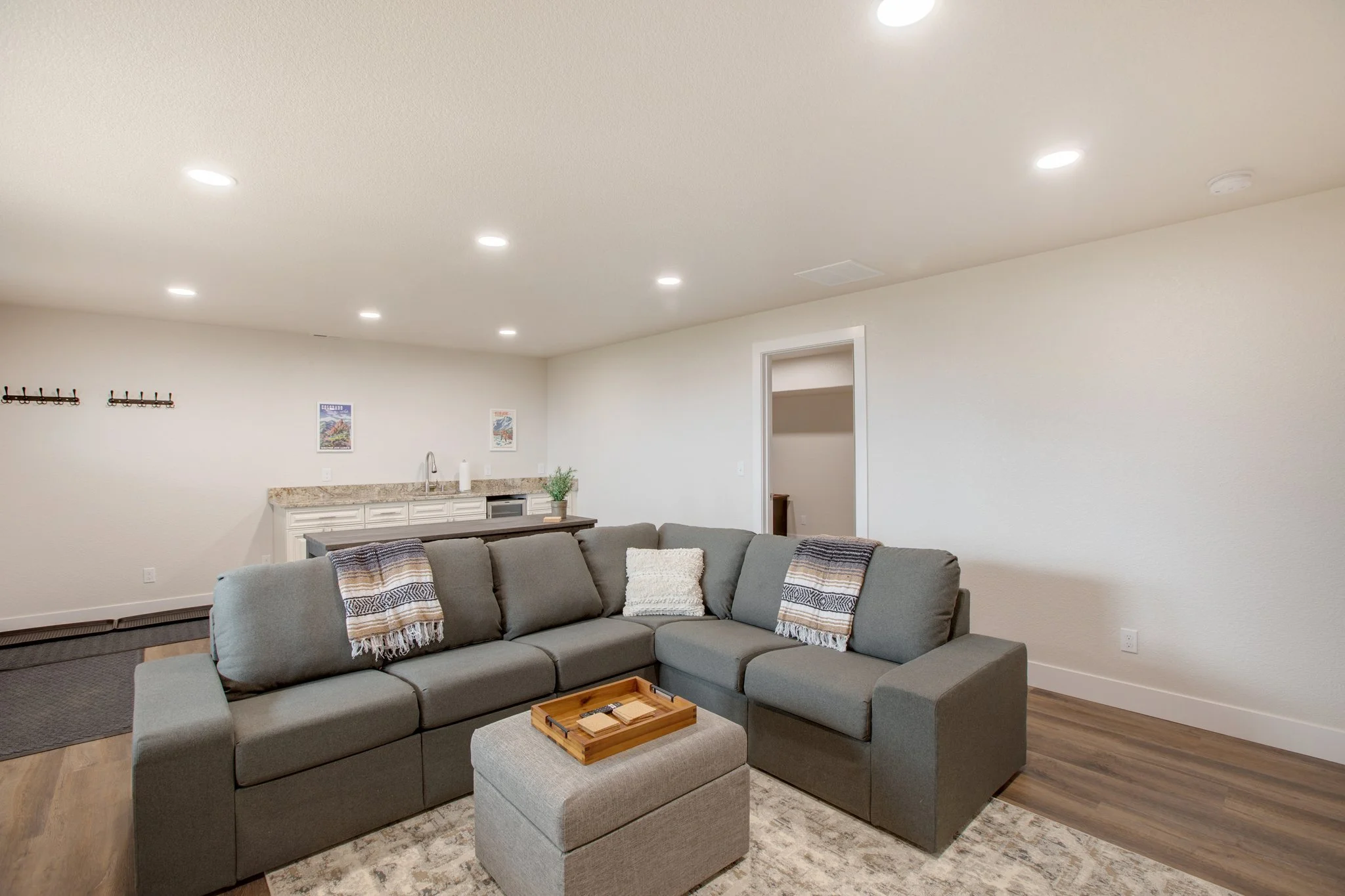 Living room with a gray sectional sofa, small matching ottoman, decorative throws, and a wooden tray on the ottoman. In the background, a small kitchenette with white cabinets, a granite countertop, and wall art. The ceiling has multiple recessed lig