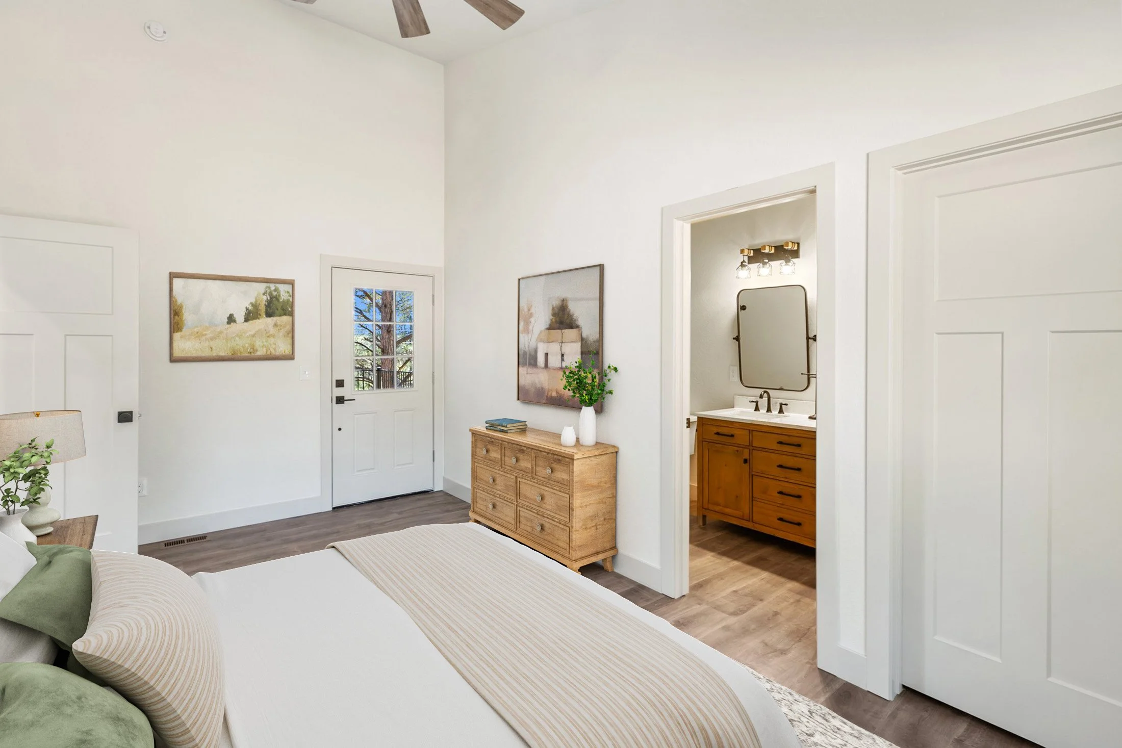 A bedroom with light wood flooring, white walls, and a bed with beige and green bedding. A wooden dresser with plant and books is against the wall, and framed landscape art hangs above it. There is a door with a small window leading outside, and an o