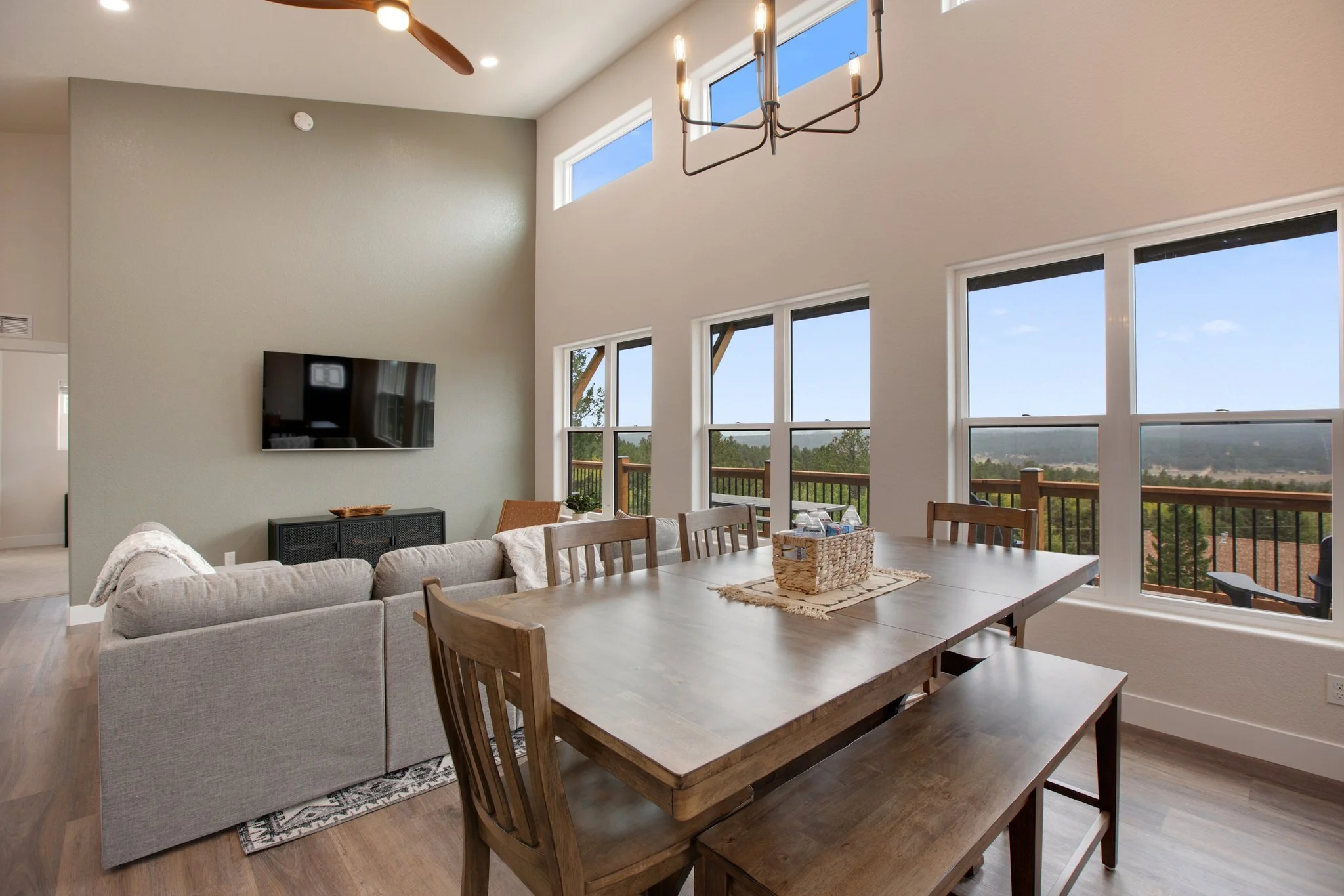 Open-concept living and dining area with large windows showing an outdoor view, a light-colored sofa, flat-screen TV on the wall, a wooden dining table with chairs, and a chandelier hanging from the ceiling.