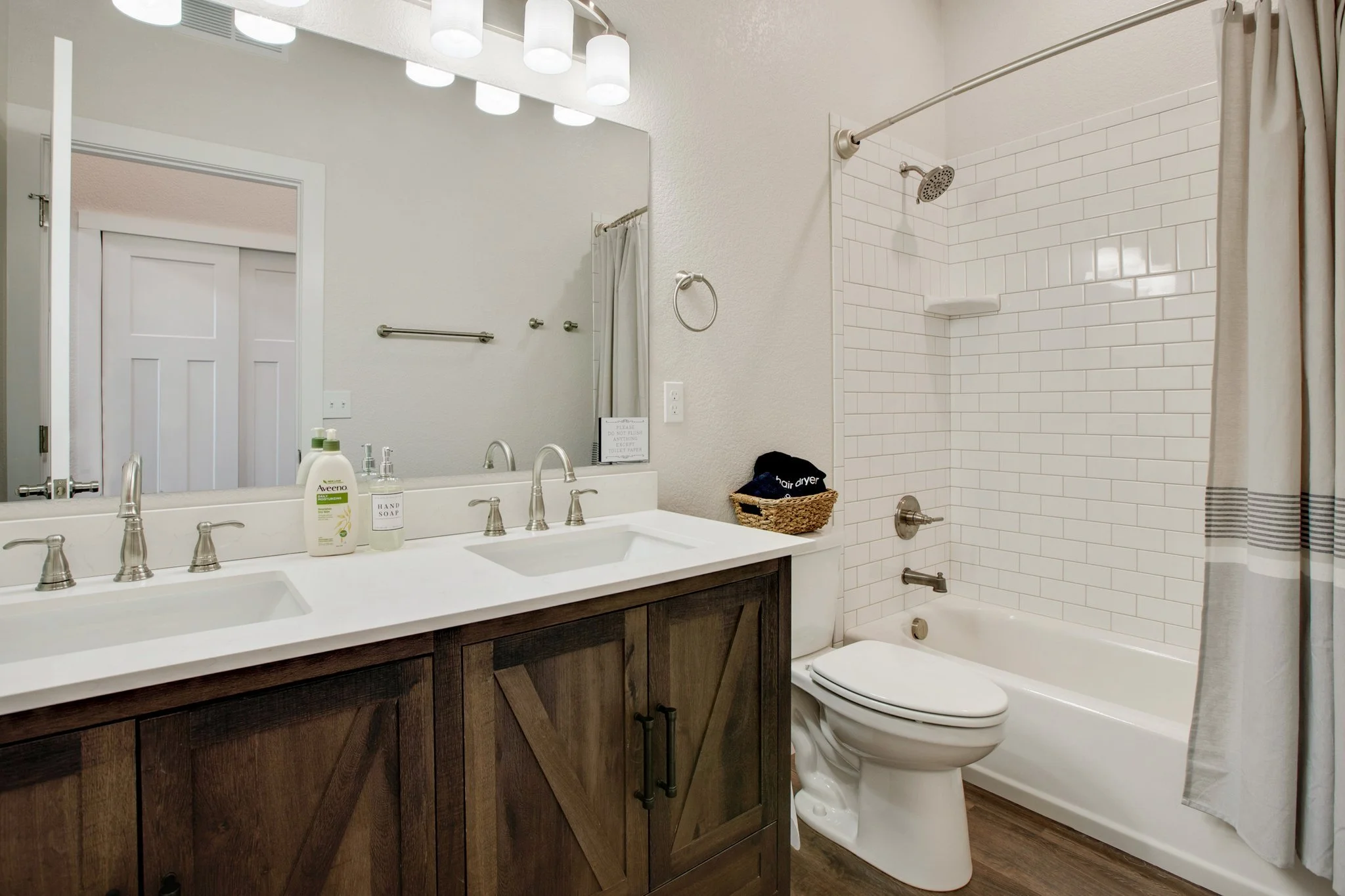 A bathroom with a dual sink vanity featuring a dark wooden cabinet, a large mirror, and modern chrome faucets. Items on the counter include a soap dispenser, hand soap, and lotion. To the right, there is a white toilet next to a shower with white sub