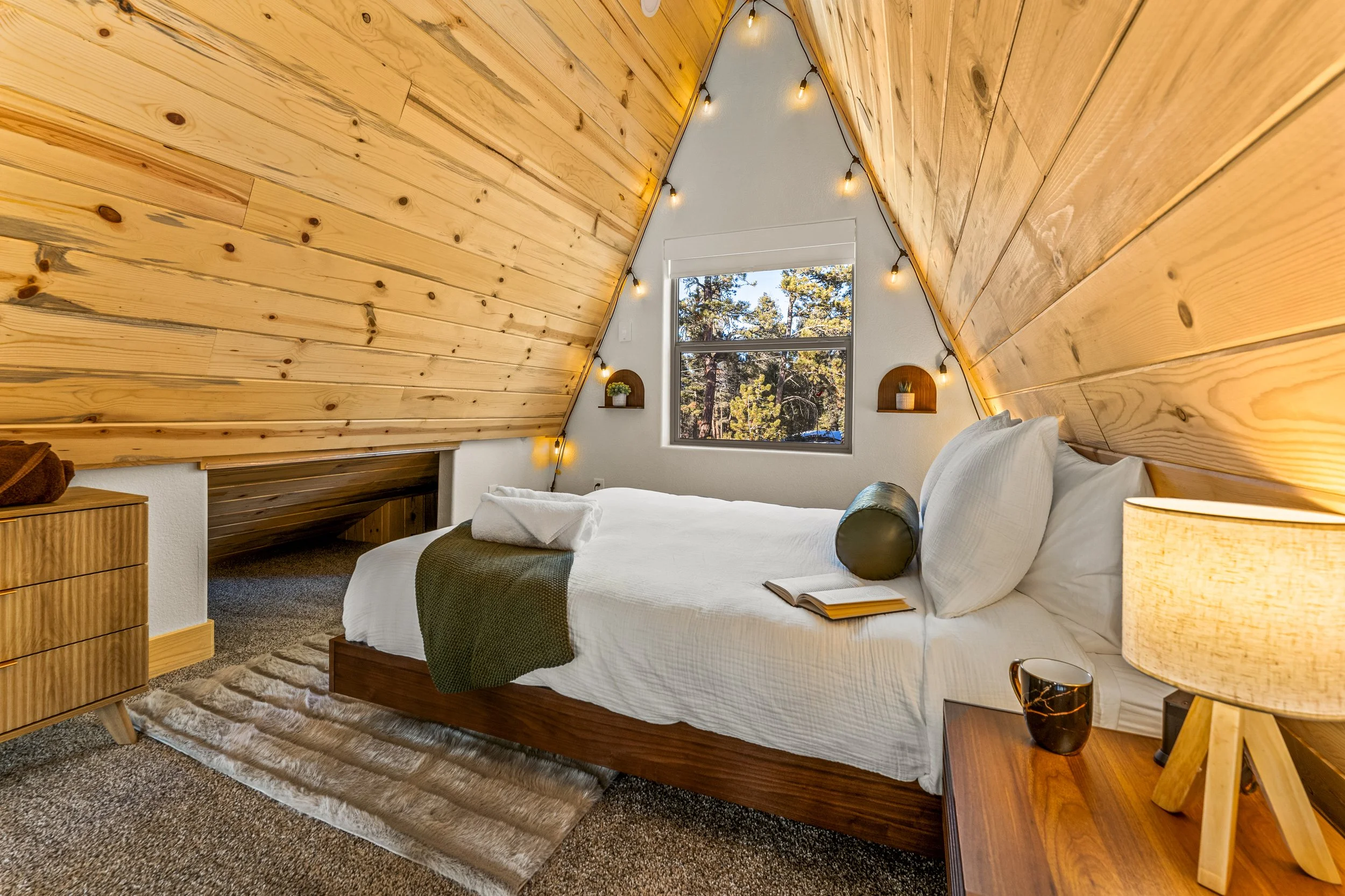 A bedroom with a wooden, triangular ceiling, a large window showing trees outside, a bed with white bedding, a bedside table with a lamp and a dark vase, and small shelves with plants and decorations.