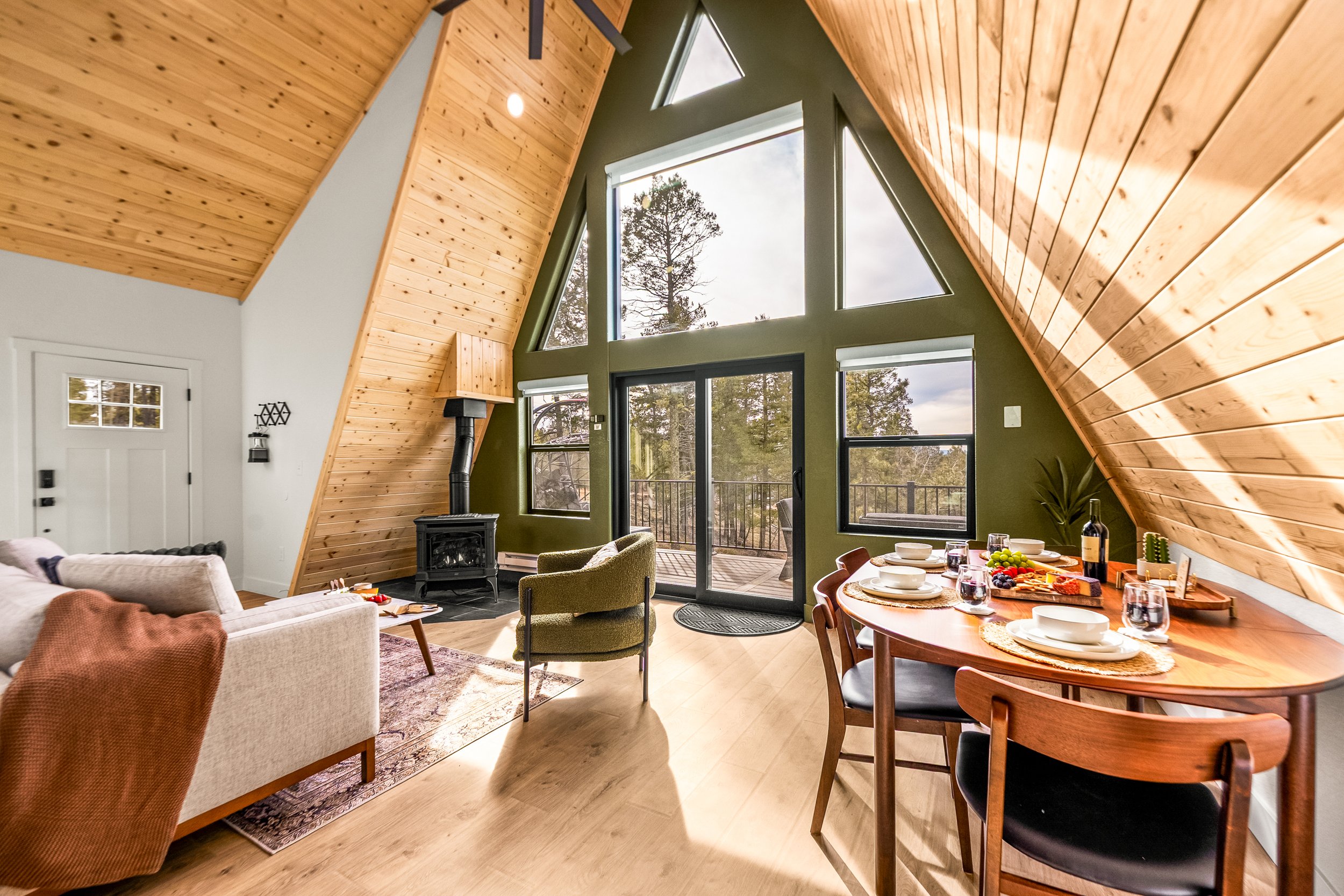 Interior of a modern home with high vaulted ceiling covered in wood paneling, large triangular and rectangular windows letting in natural light, and a cozy living and dining area with furniture and table settings.
