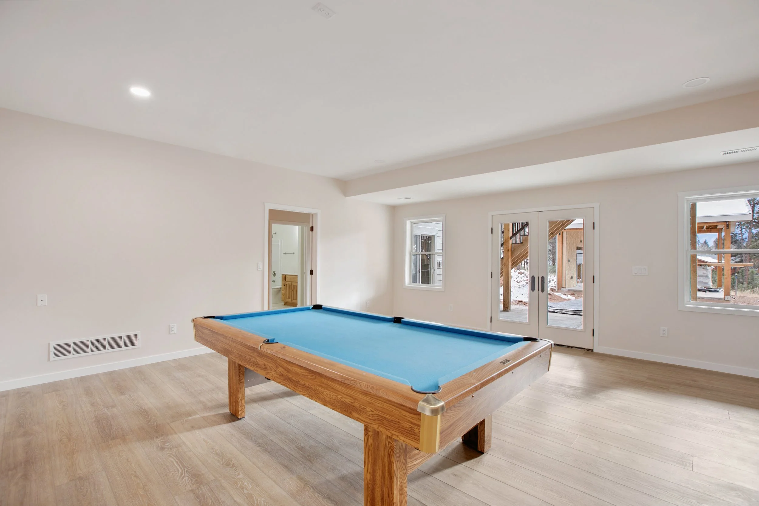 A spacious, unfinished room with light-colored walls and wooden flooring, featuring a pool table with a blue felt surface. Large windows and glass double doors provide natural light and show an outdoor construction area with wooden beams and snow, in