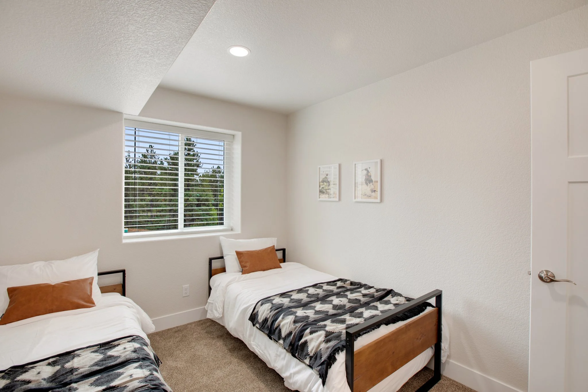 A bedroom with white walls and a window with blinds overlooking trees. Two beds with white linens, brown and white pillows, and black-and-white patterned blankets.
