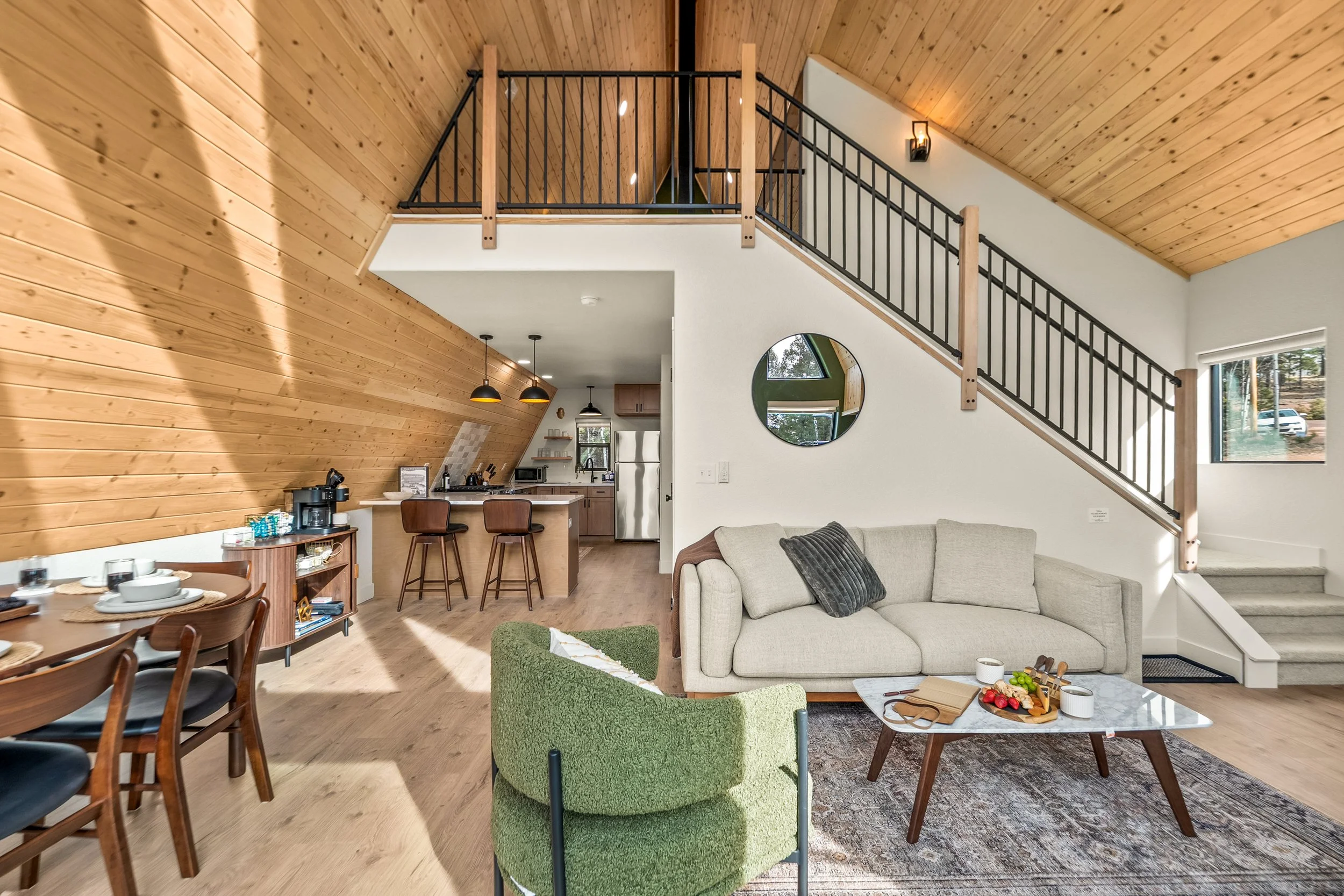 Interior of a living room with wooden ceiling, white walls, and large windows. Features include a beige sofa with pillows, a round marble coffee table with snacks and cups, a green armchair, and a kitchen area with a breakfast bar and bar stools.