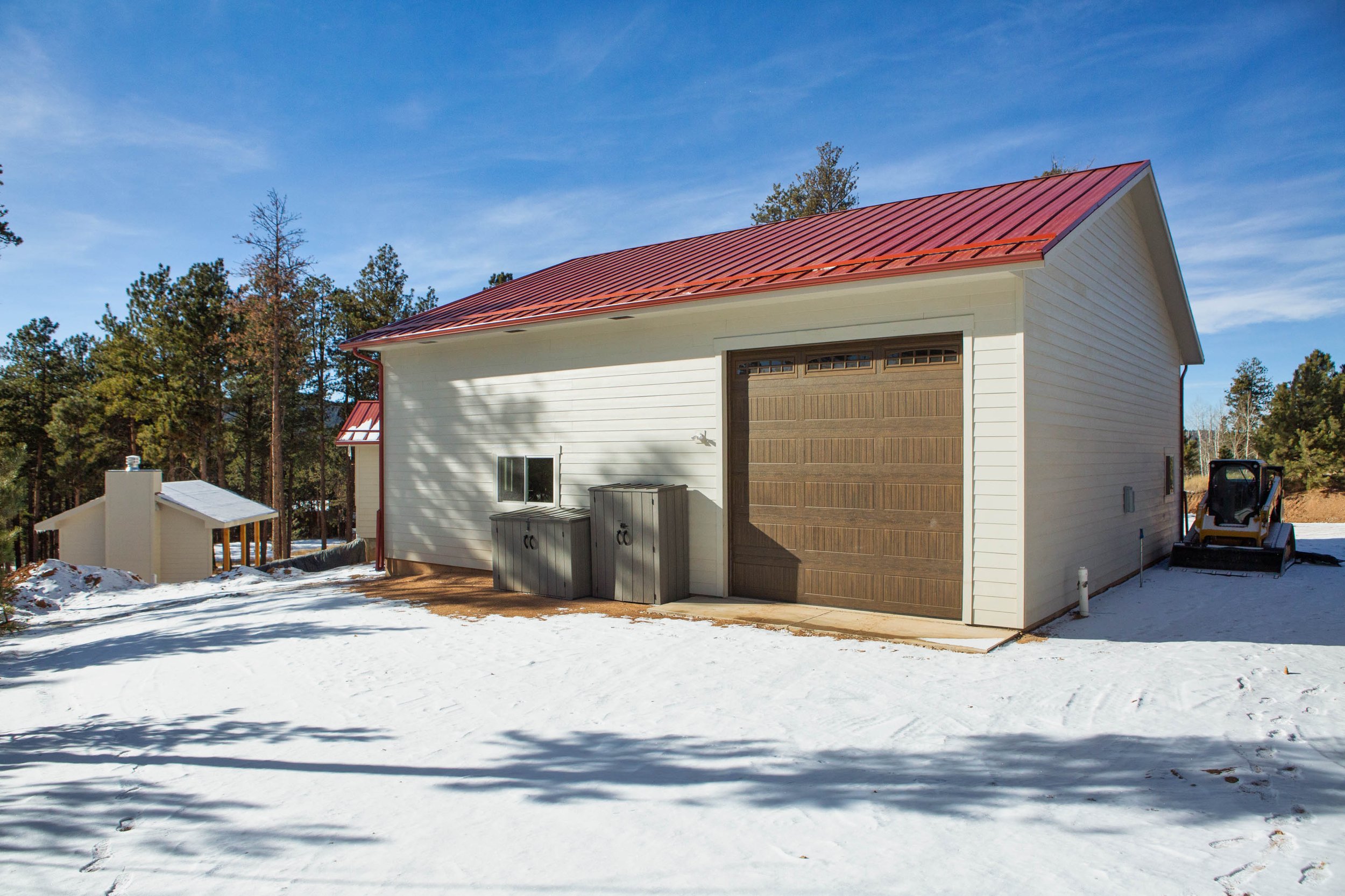 A white house with a red metal roof, brown garage door, and small window, surrounded by snow and trees under a blue sky.