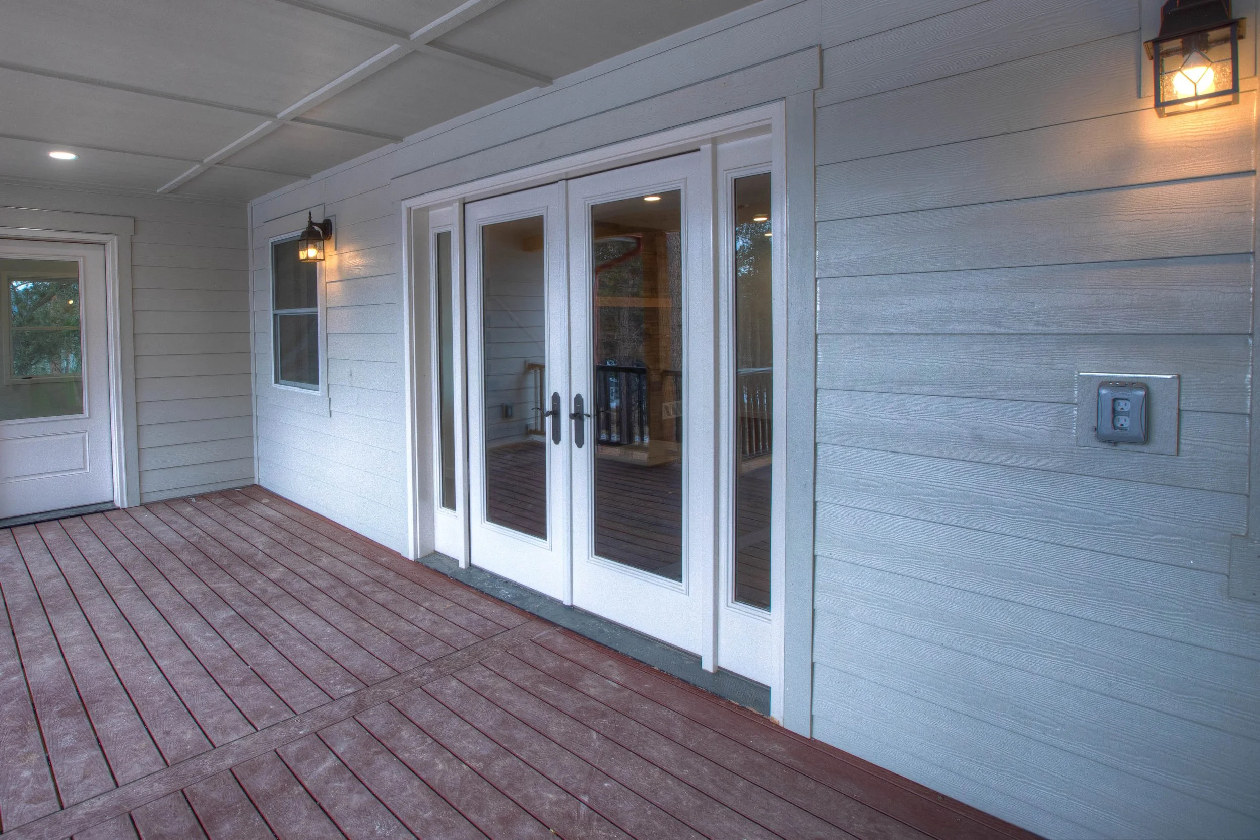Exterior porch with sliding glass doors, a side window, and wall-mounted light fixtures on a gray house with wood siding and a red wood deck.