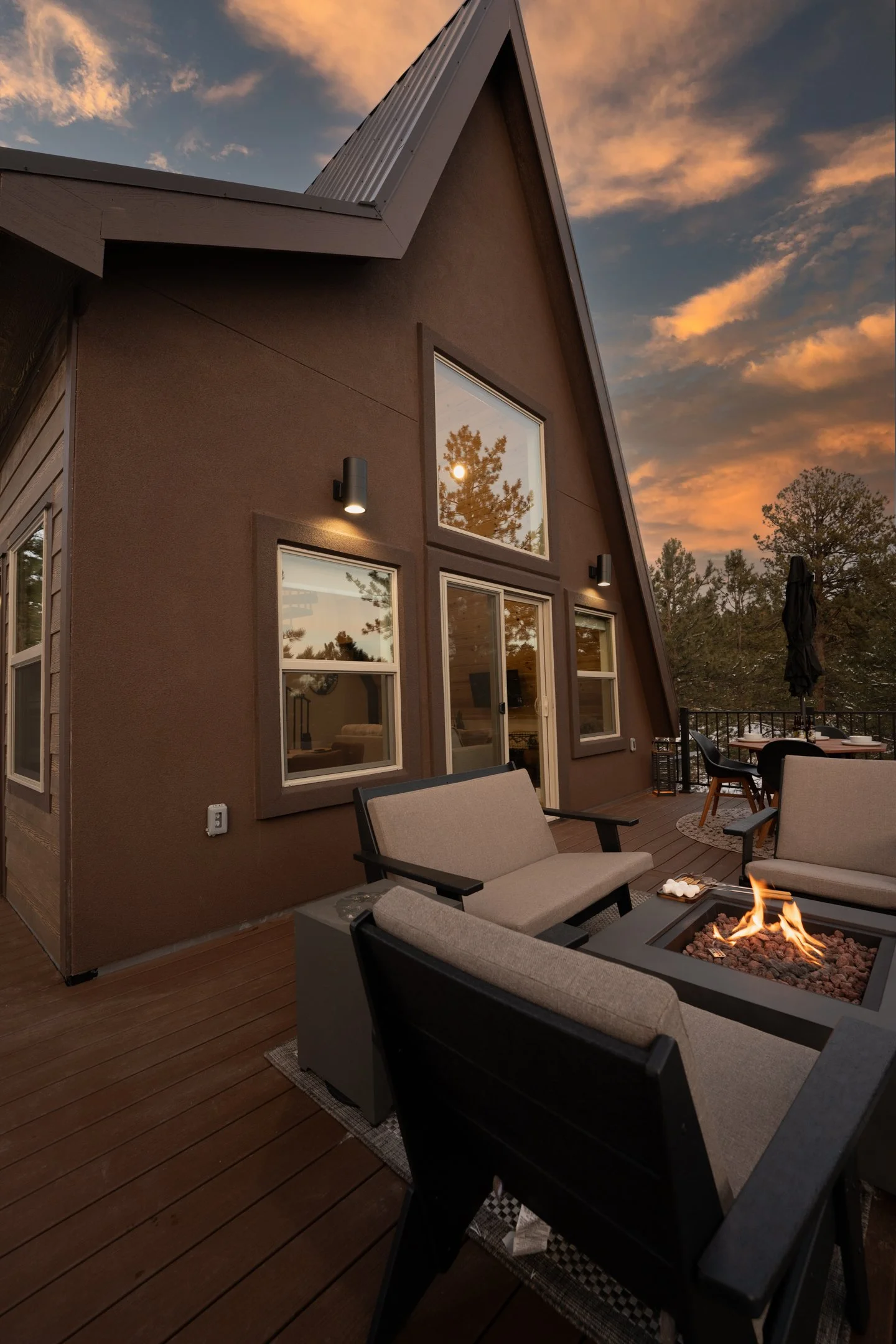 A backyard outdoor patio area features beige cushioned chairs surrounding a fire pit with a small flame, with a brown house wall in the background and a sunset sky with orange and blue clouds.