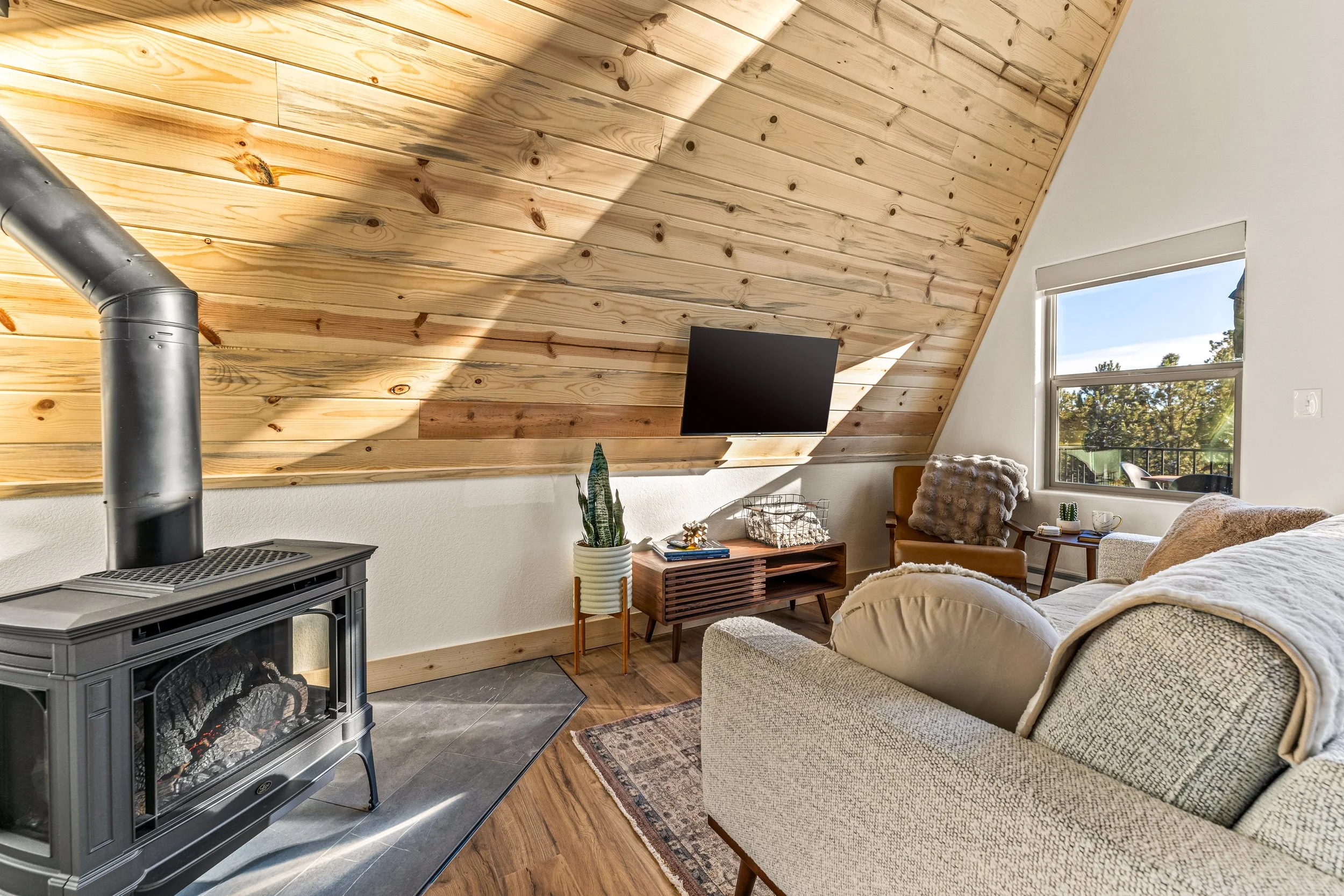Living room with angled wood-paneled ceiling, large window, sofa, armchair, fireplace, and a potted plant.