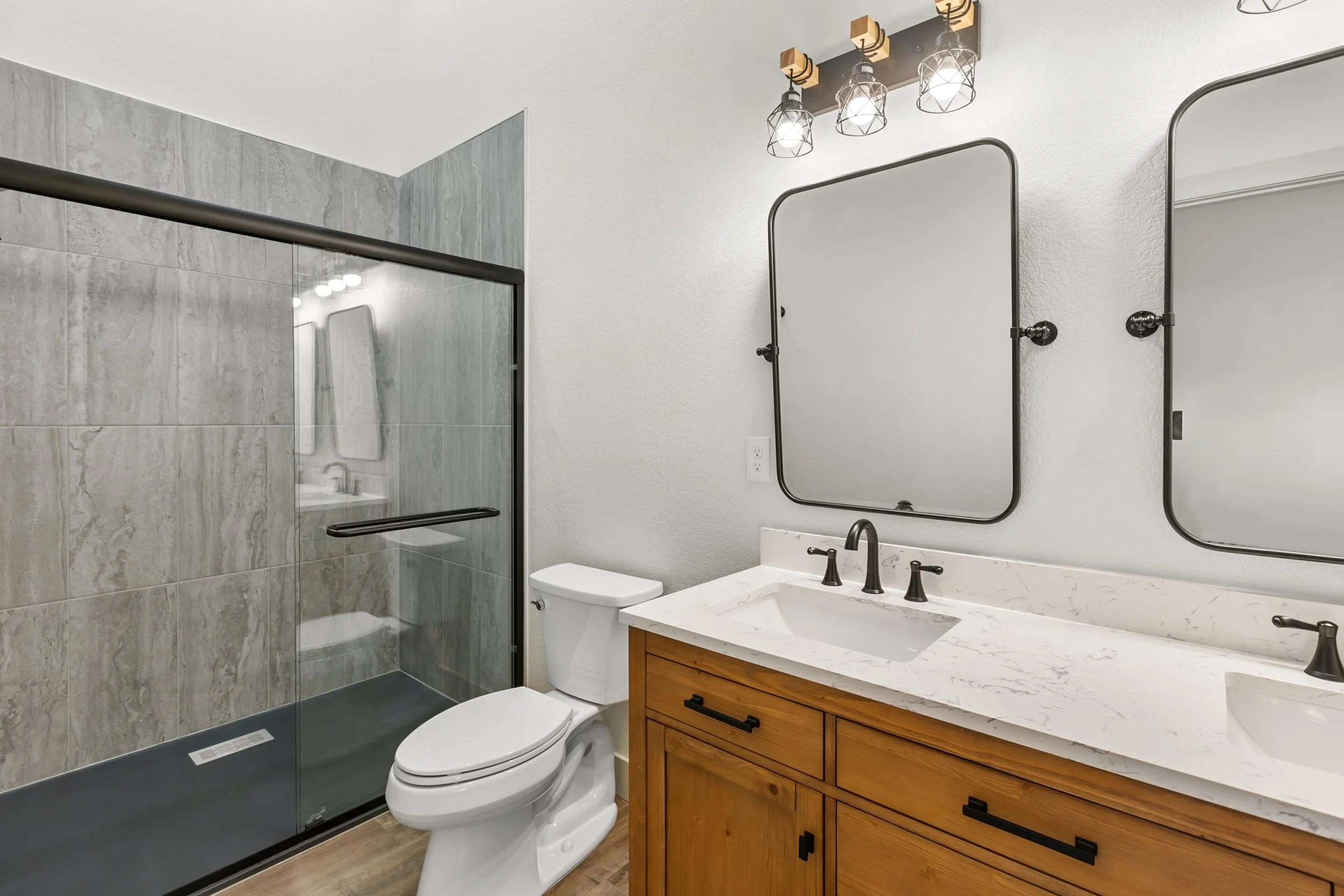 Modern bathroom with a walk-in shower with glass door, white walls, wood vanity, marble countertop, large mirrors, and industrial style lighting fixtures