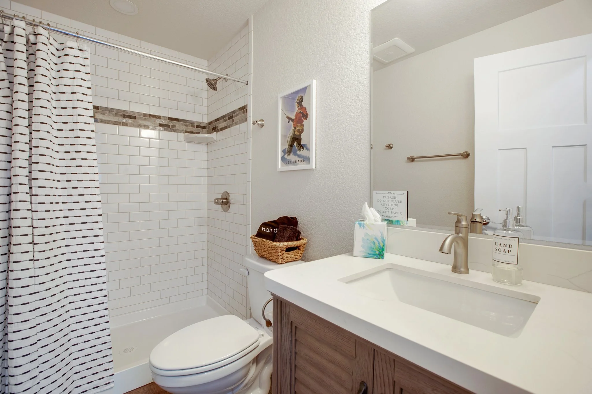 A bathroom with a shower area enclosed by a curtain, white tiled walls with a decorative strip, a toilet, a vanity with a sink and a large mirror, and various toiletries on the counter.