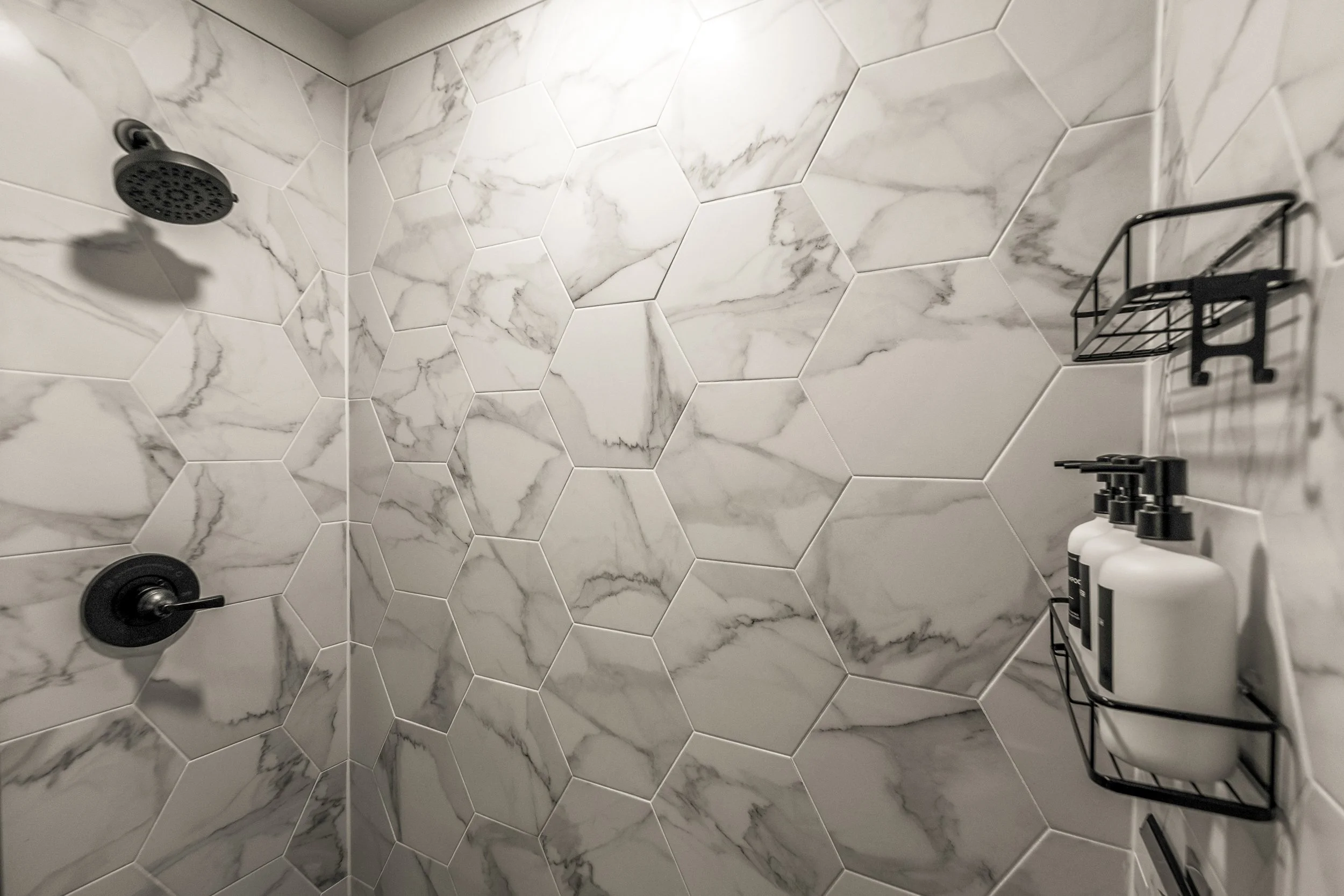 A bathroom shower area with white hexagonal tiles with gray veining on the walls. There is a black showerhead on the upper left side, a black handle and control on the lower left, and a black metal shower caddy on the right holding three white bottle