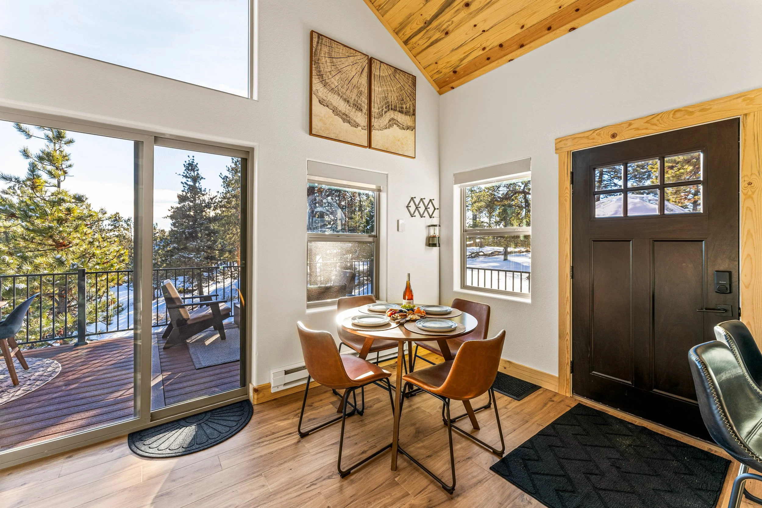 A dining area with a round table set with plates and pretzels, surrounded by six tan chairs, with large windows showing snow-covered trees outside. An outdoor porch with chairs and a railing is visible through the sliding glass door.