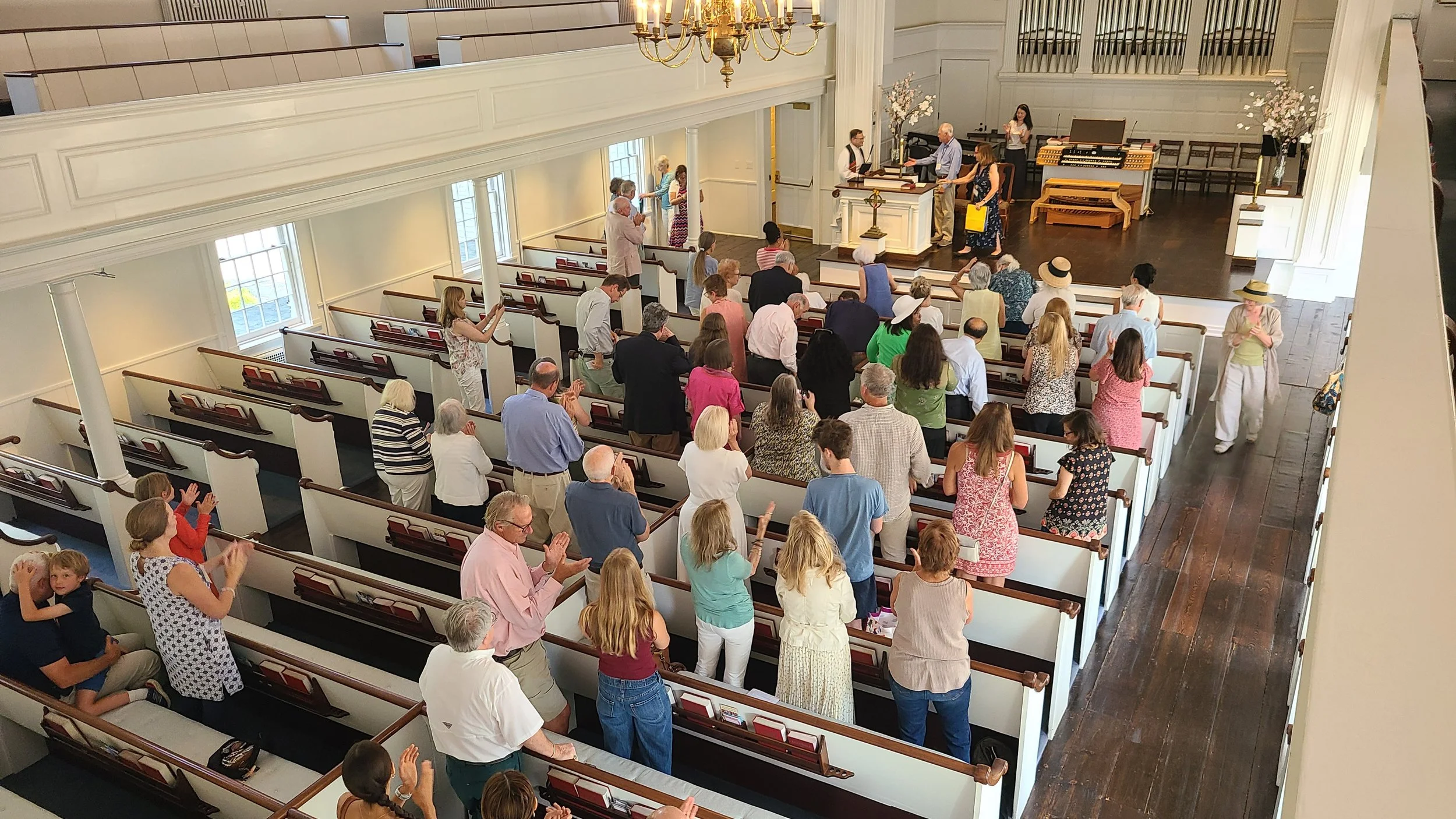A congregation of people gathered inside a church, standing and praying during a service. Some individuals are clapping, others are kneeling, and a few are taking photos. The church interior features white walls, wooden pews, large windows, a wooden stage with a musical organ, and floral arrangements. A man is at the pulpit, and several people are near the altar.